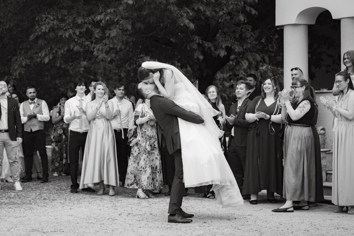 Black and white photo of a bride and groom sharing a romantic dip kiss surrounded by applauding wedding guests outdoors.