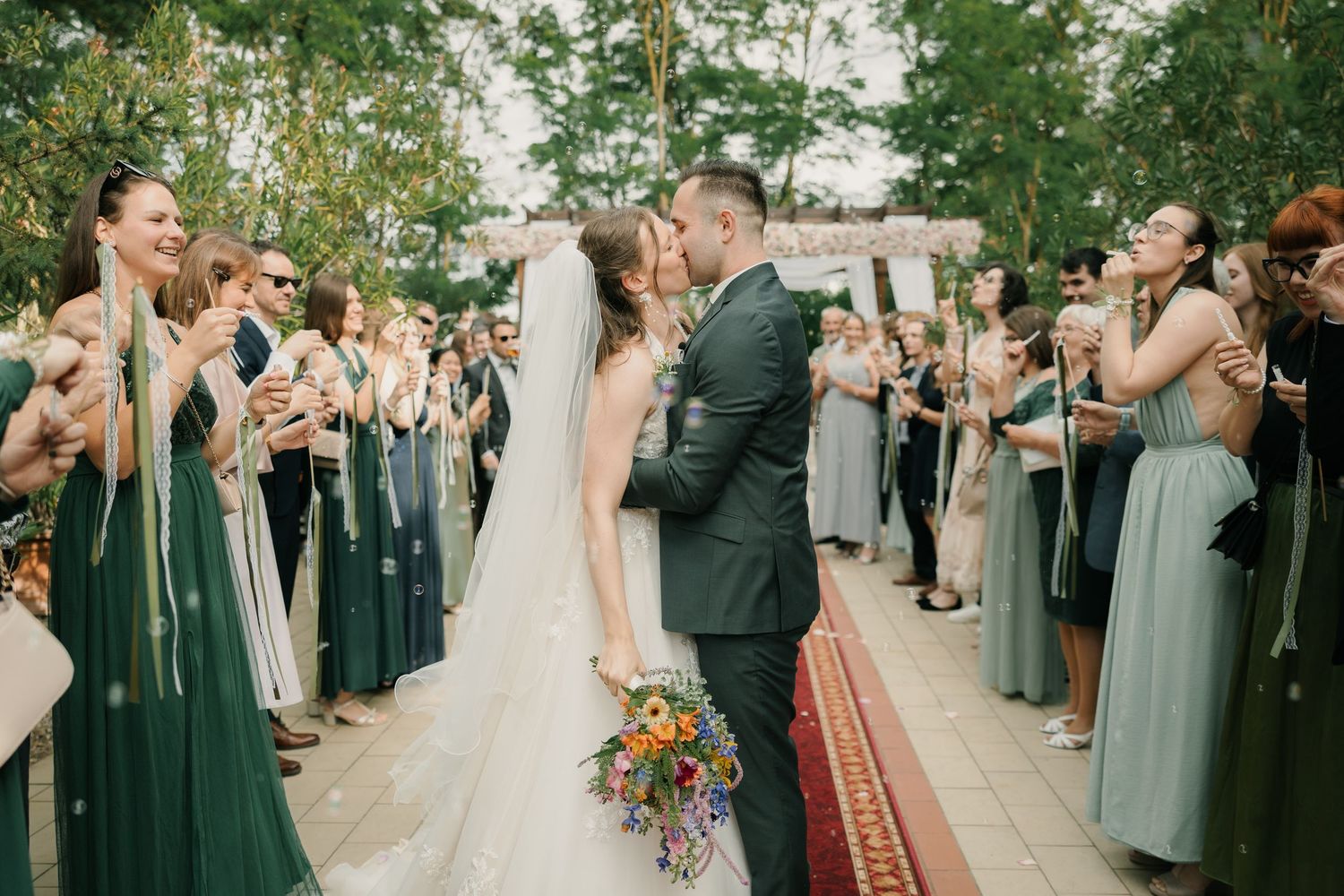 Bride and groom share a kiss at outdoor wedding ceremony, surrounded by guests and bridesmaids in green dresses.