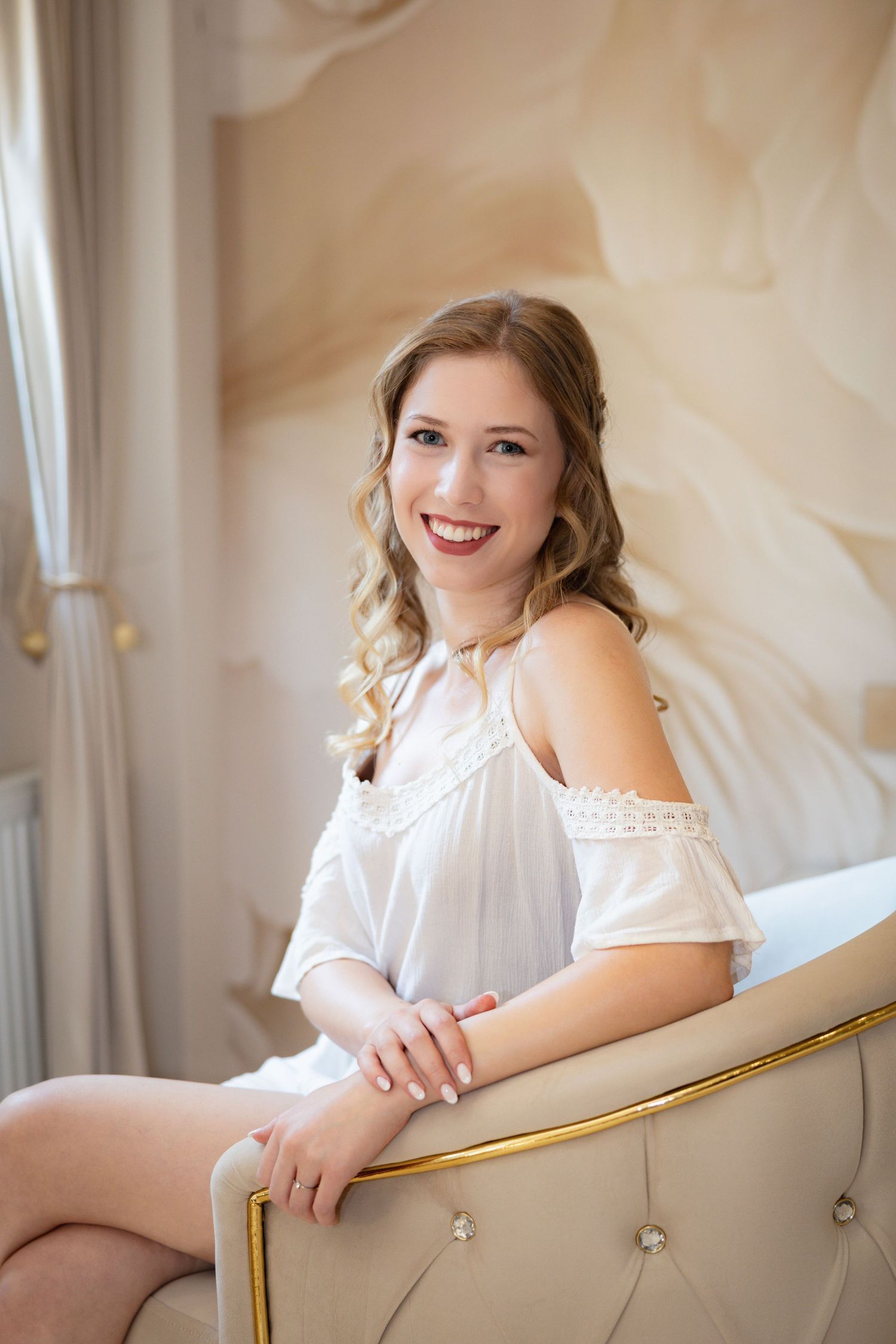 Young woman in white off-shoulder top smiling while seated in elegant beige armchair in a bright, stylish room.