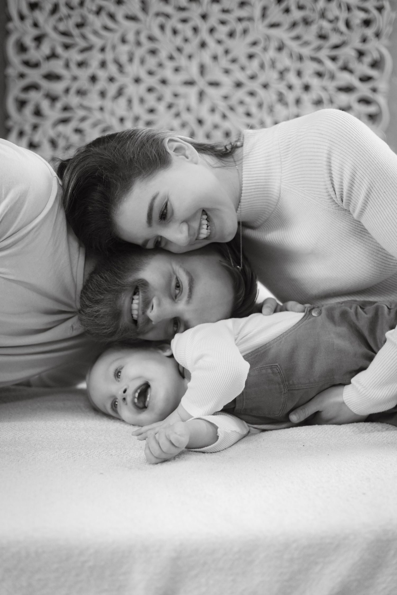 Black and white photo of happy parents smiling with their laughing baby lying on a bed.