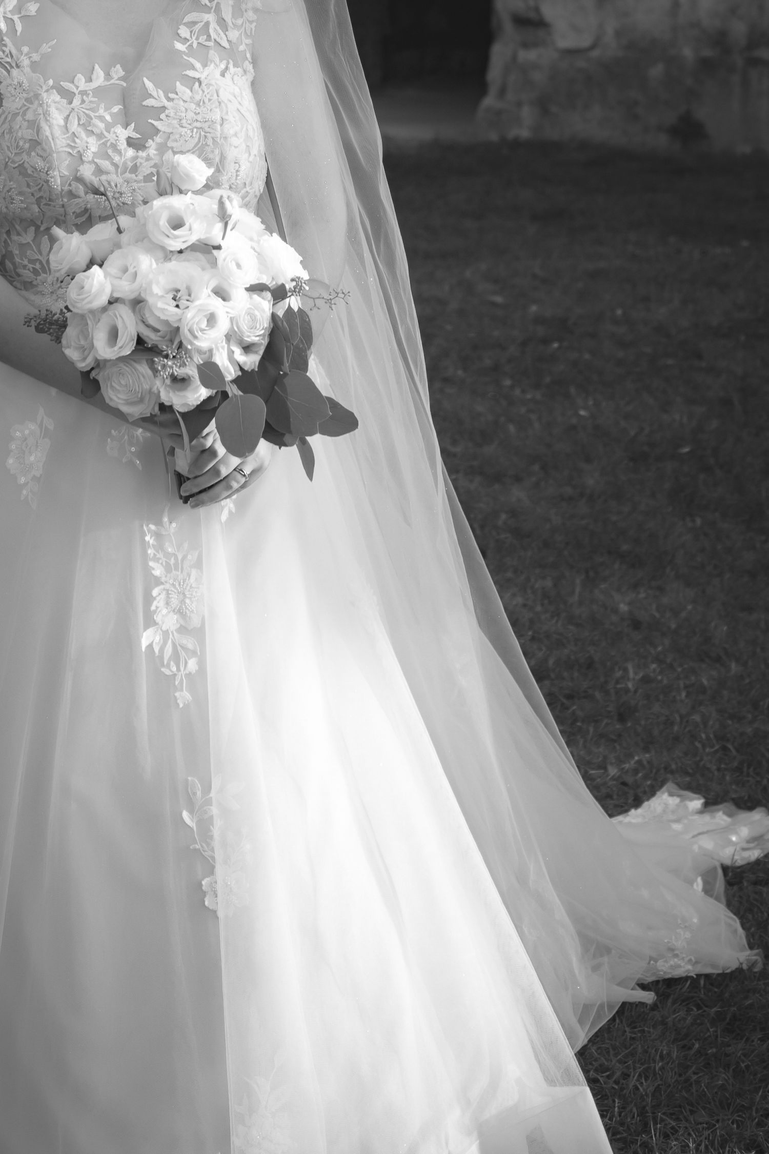A black and white photograph of a wedding bouquet held against a flowing white wedding dress and veil.