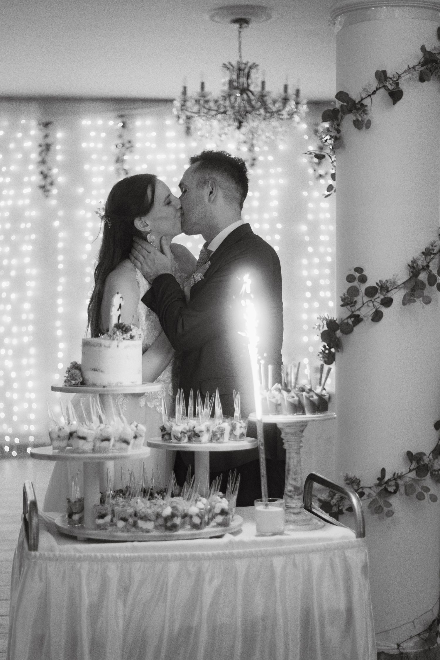 Black and white photo of a bride and groom kissing behind a wedding dessert table with cake and treats.