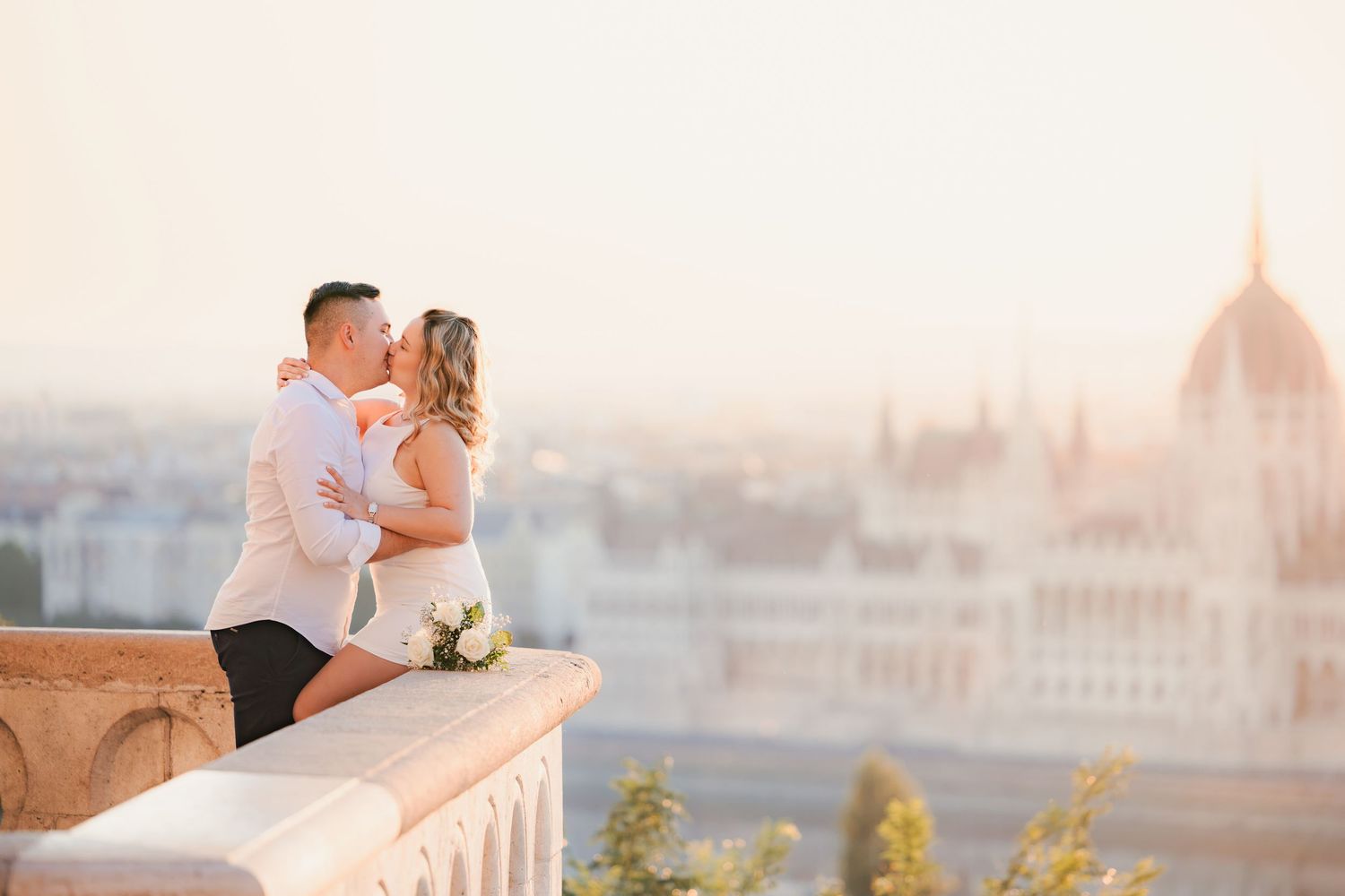 Couple sharing a romantic kiss on a stone balcony with the Hungarian Parliament Building at sunset in Budapest.