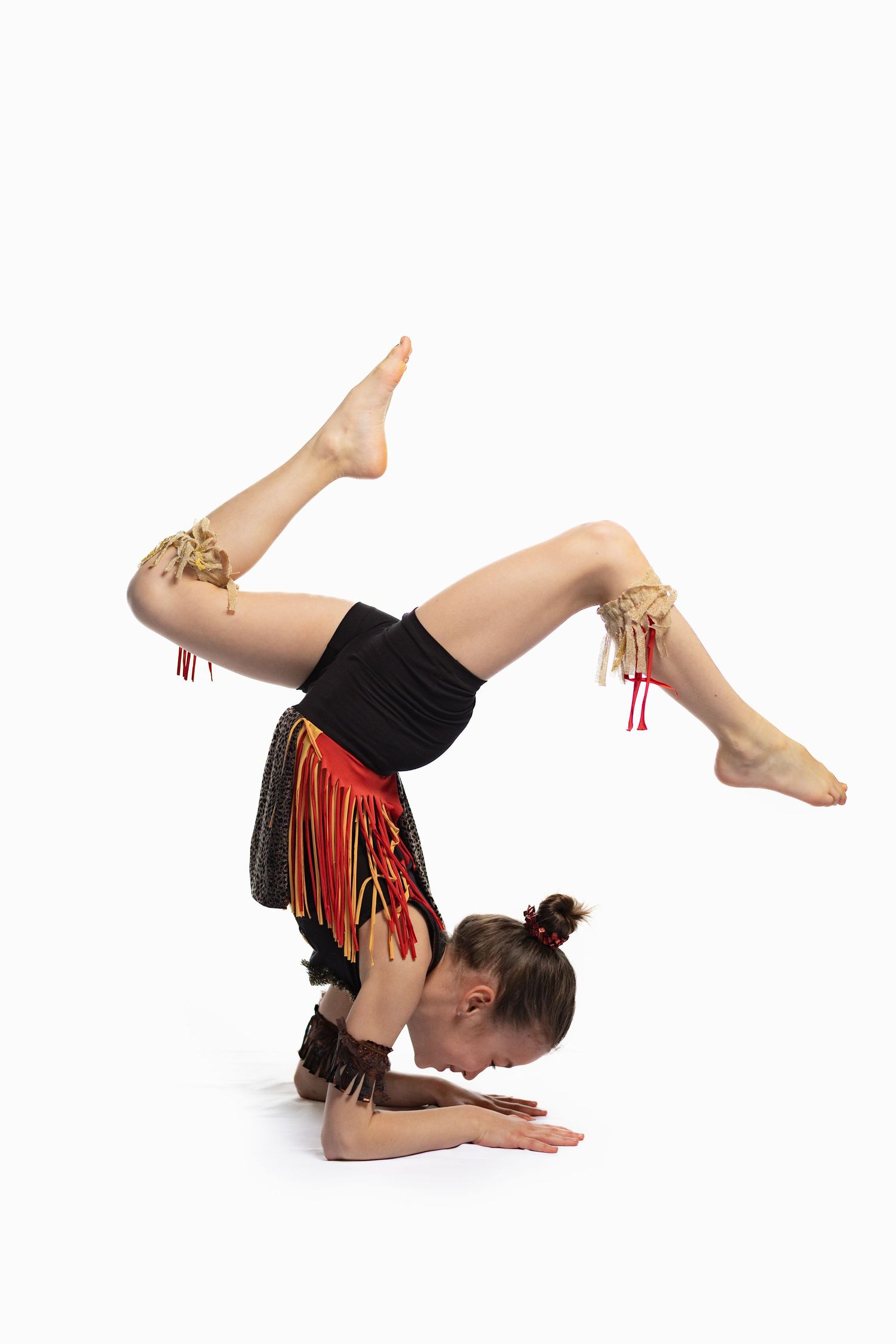 Young acrobat performing a headstand with legs bent, wearing a colorful fringed costume on a white background.