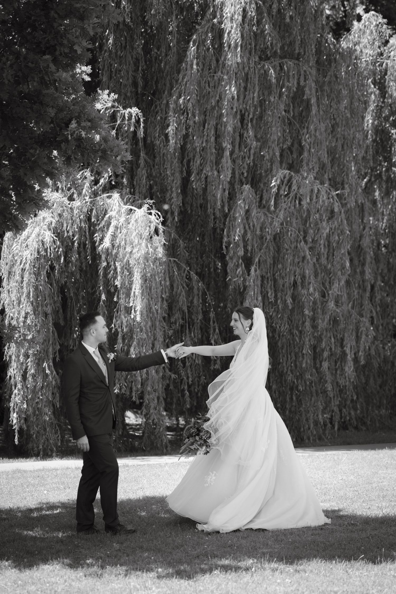 Black and white photo of bride and groom holding hands outdoors near a weeping willow tree on their wedding day.