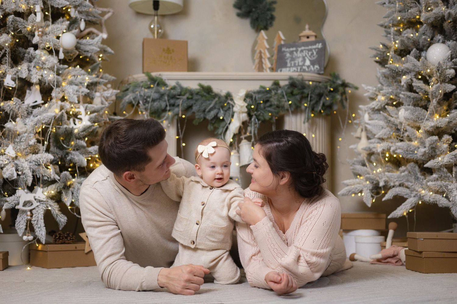 Young family with baby girl celebrating Christmas beside decorated white trees with warm lights and gifts.