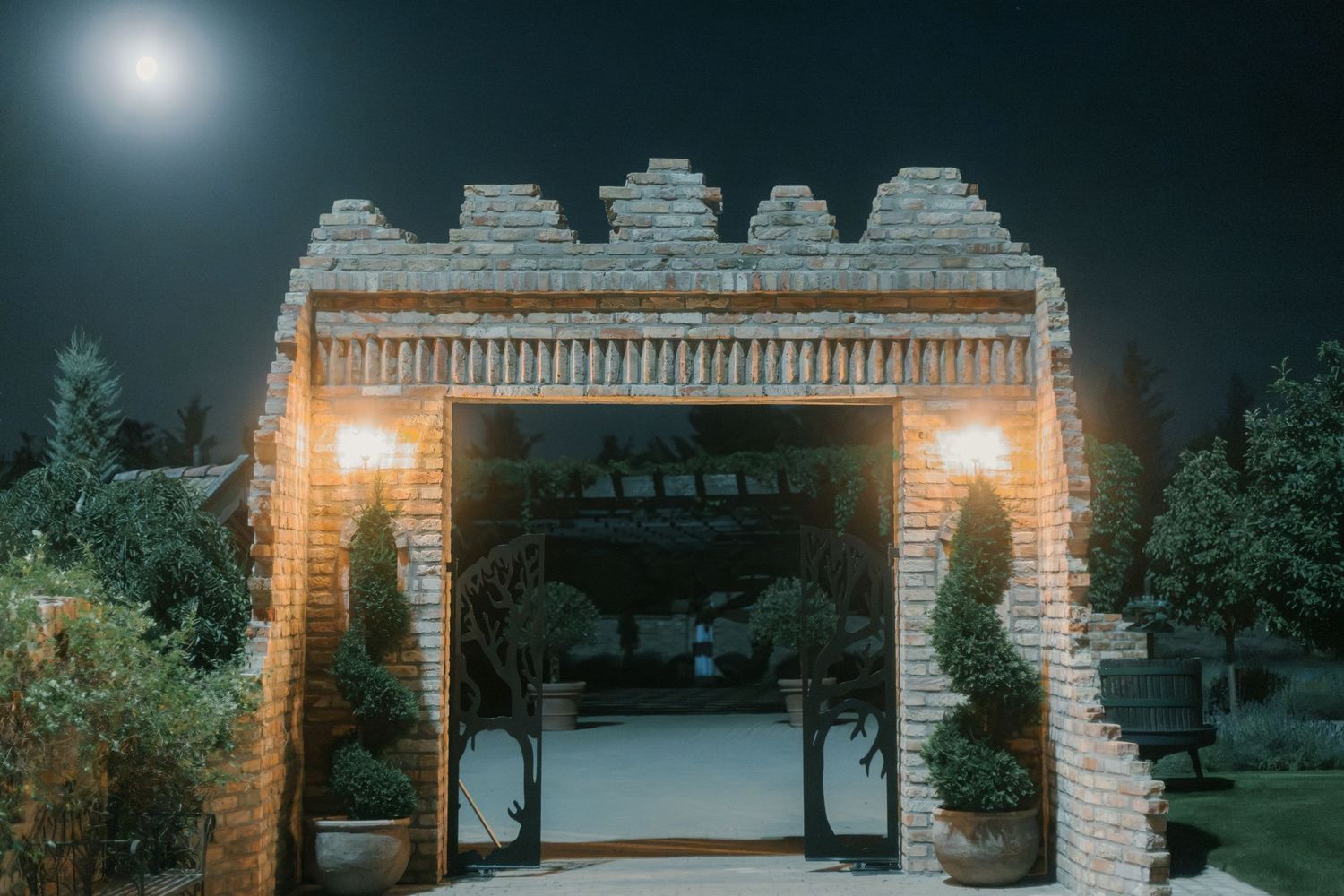 Illuminated brick archway gate at night with warm lanterns and topiary trees under a full moon.