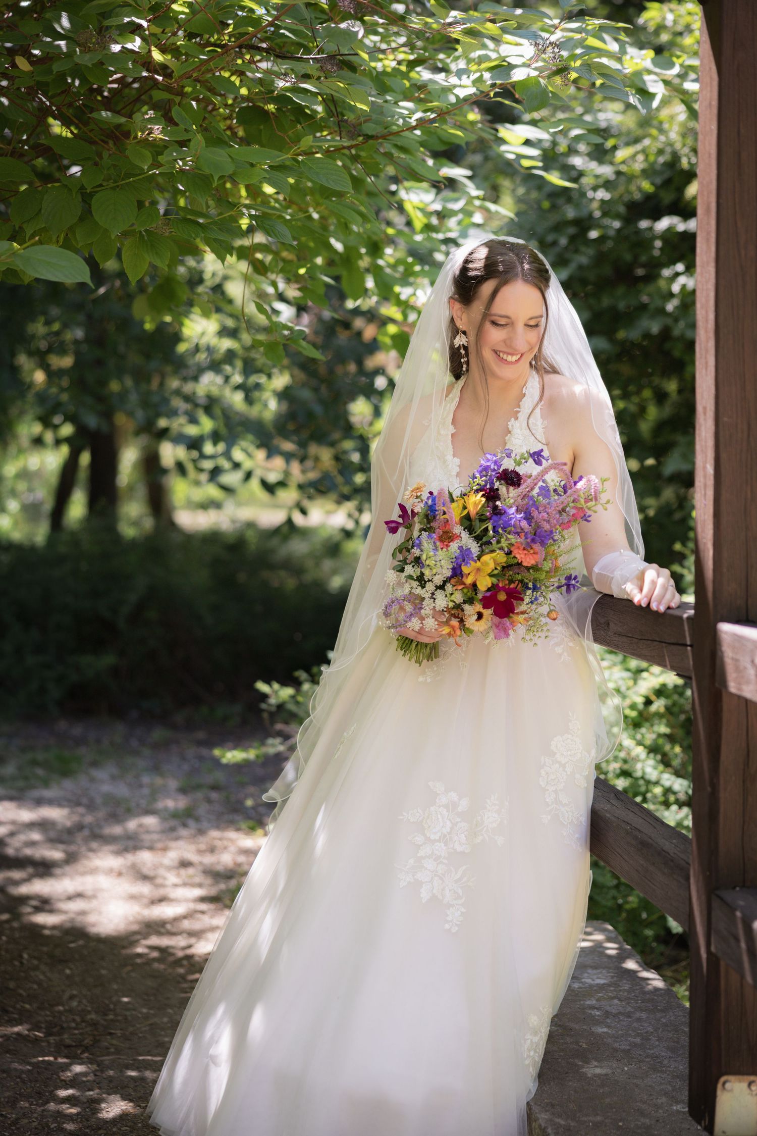 Smiling bride in white lace gown and veil holds colorful wildflower bouquet on garden steps surrounded by lush greenery.