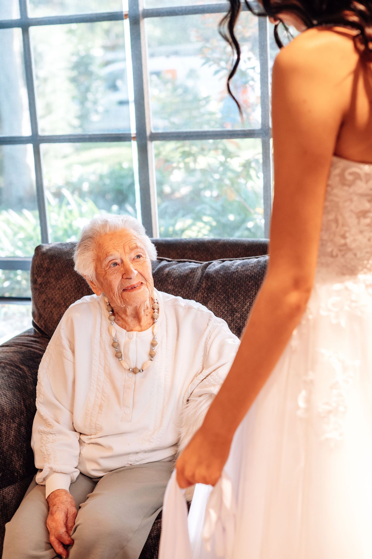 Elderly woman in white outfit gazes up at bride in lace wedding gown during an emotional pre-wedding moment.