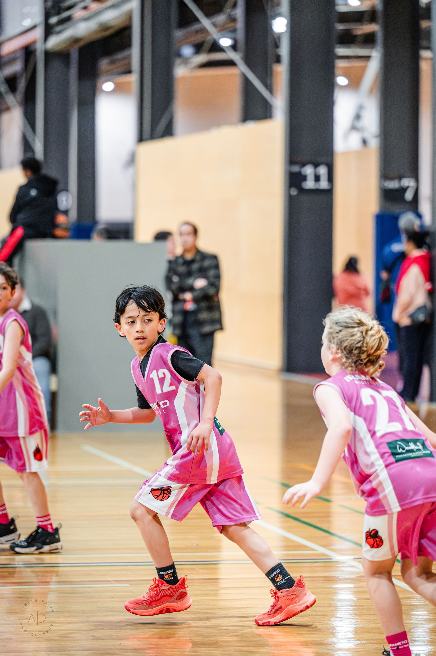 Young children in pink basketball uniforms playing an indoor youth basketball game on a hardwood court.