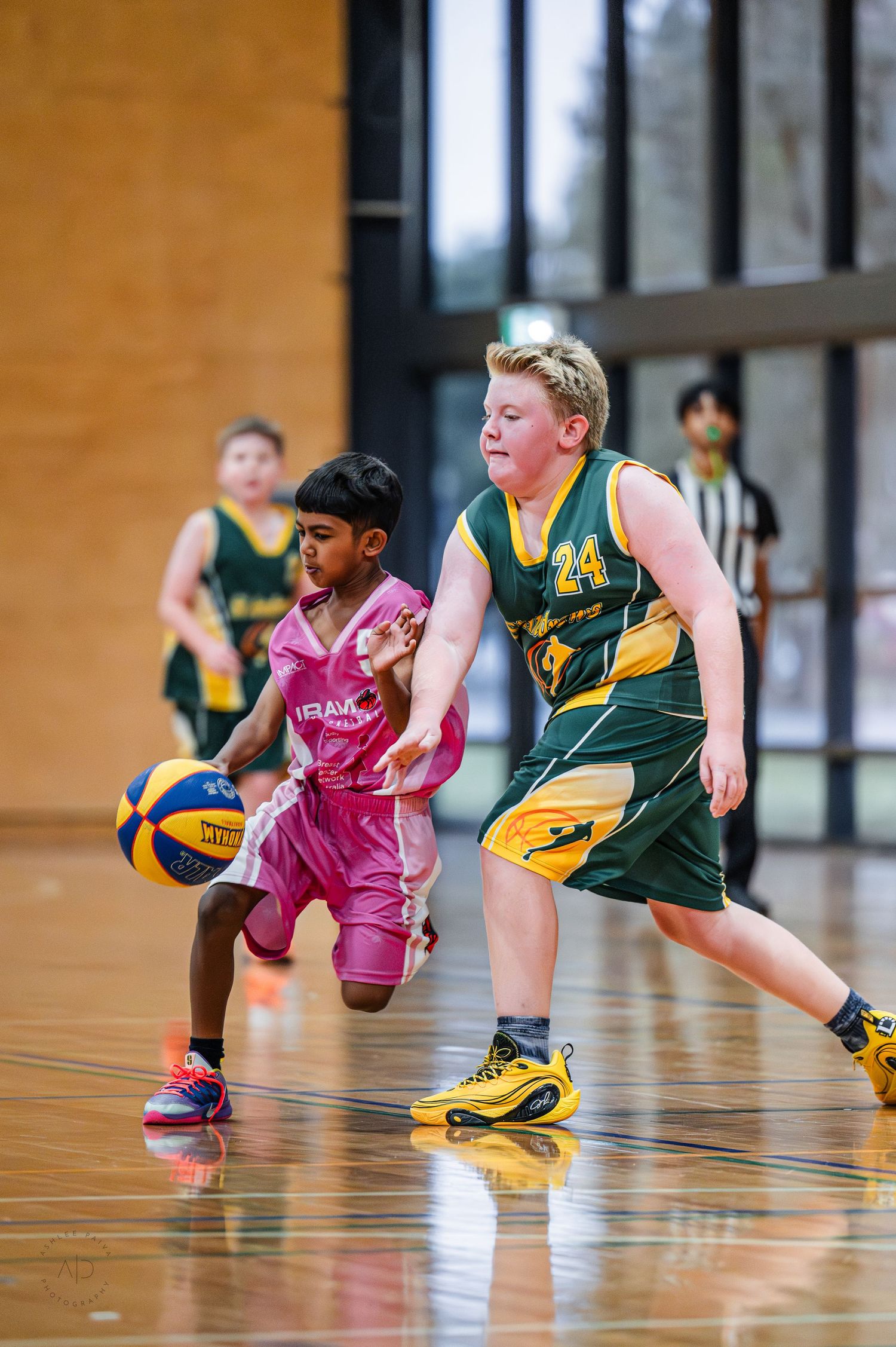 Two youth basketball players compete on indoor court, player in pink dribbles past defender in green number 24 jersey.