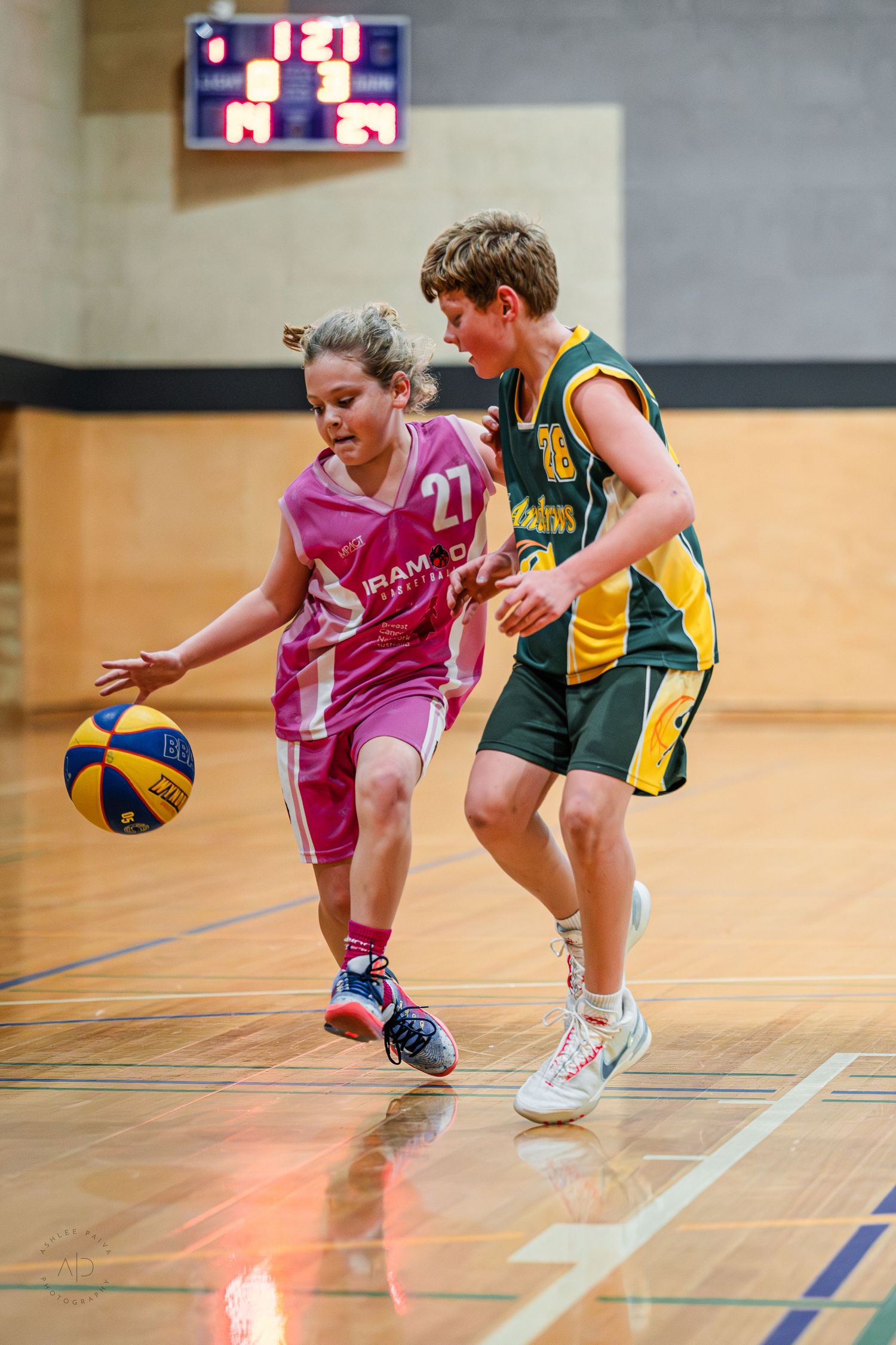 Two youth basketball players compete on indoor court, child in pink jersey #27 dribbling past green and gold defender.