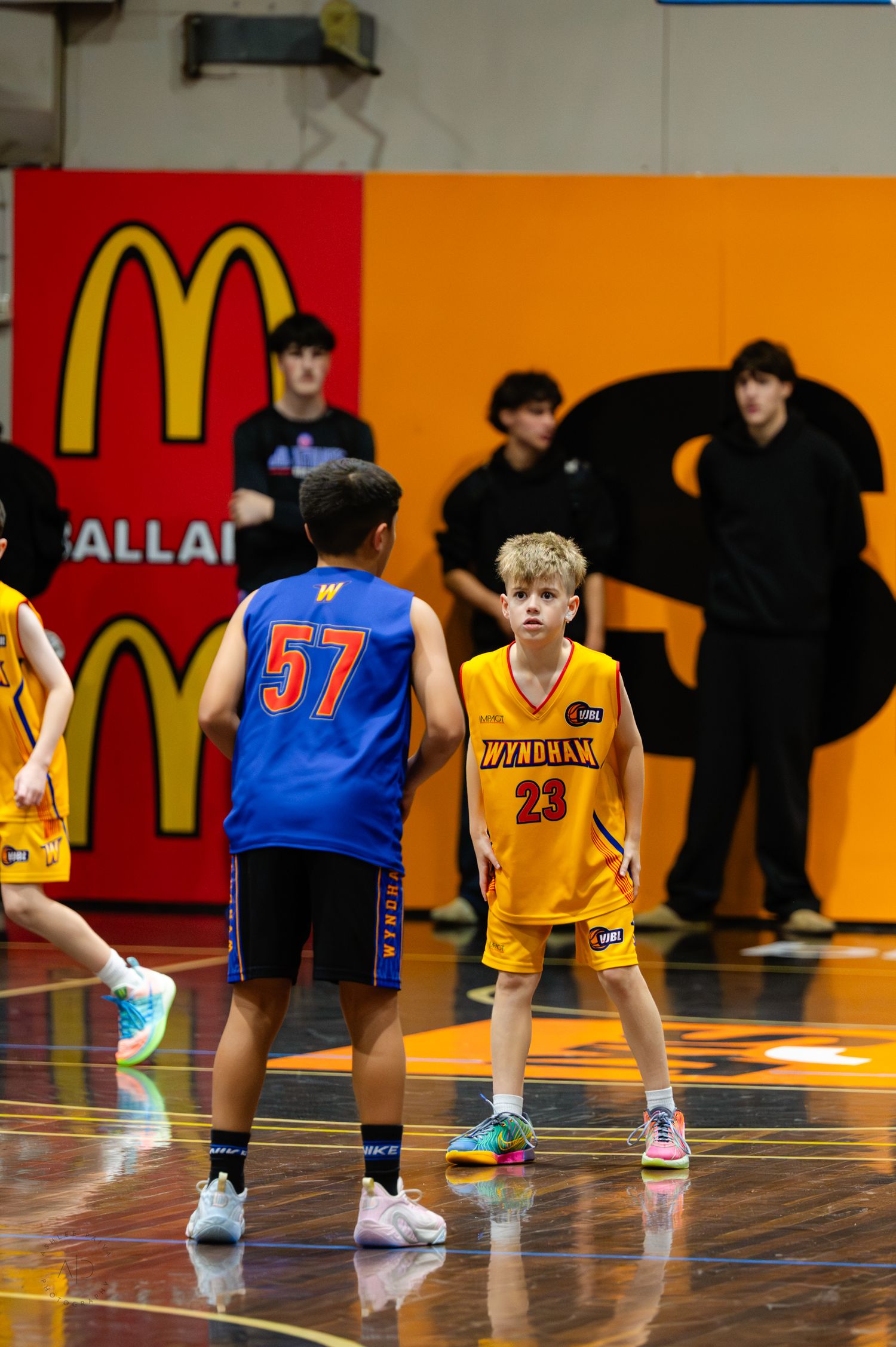 Two youth basketball players in blue #57 and gold #23 Wingham jerseys face off on an indoor court with McDonald's signage.