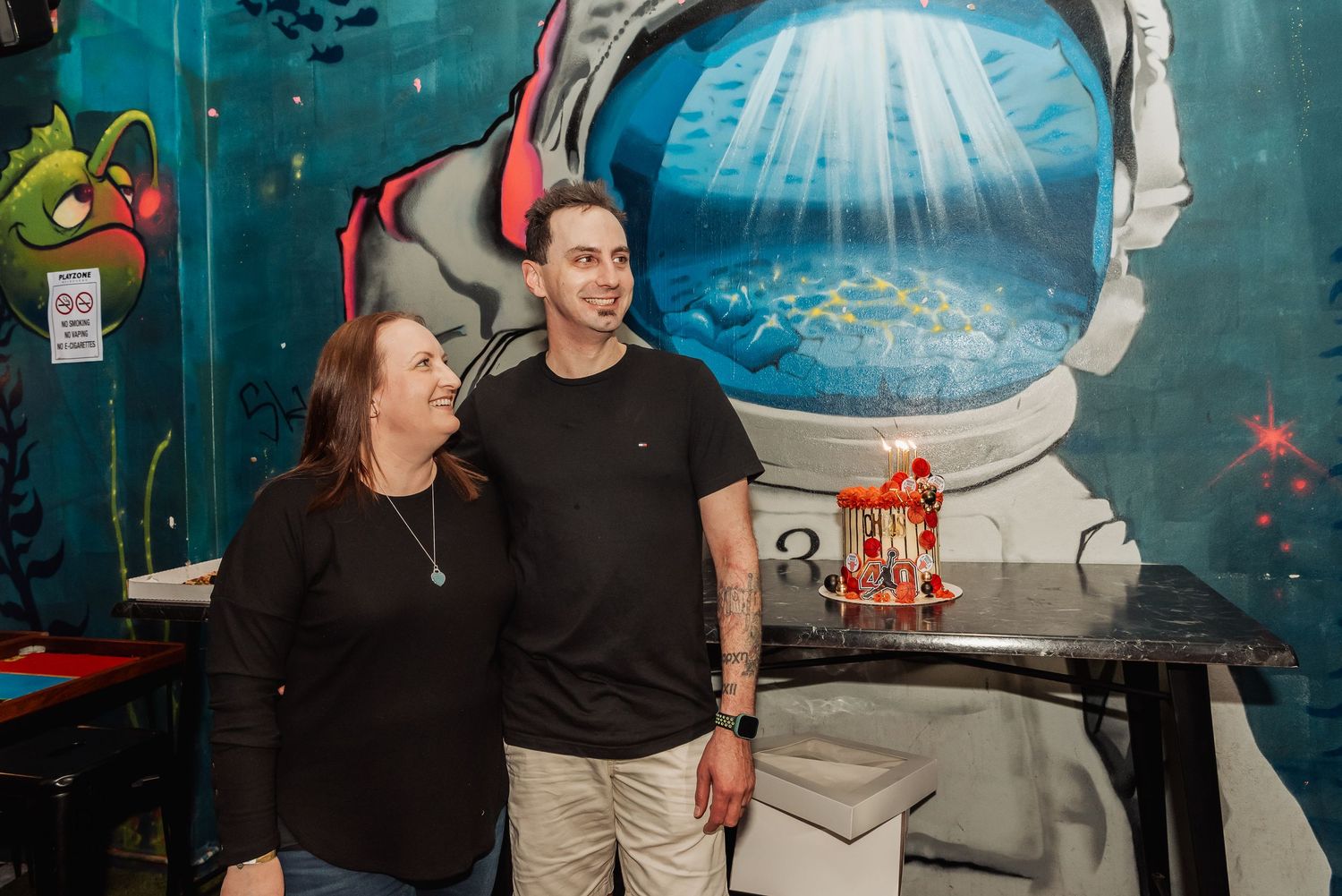 Two people smiling in front of a colorful astronaut mural in a vibrant restaurant setting.