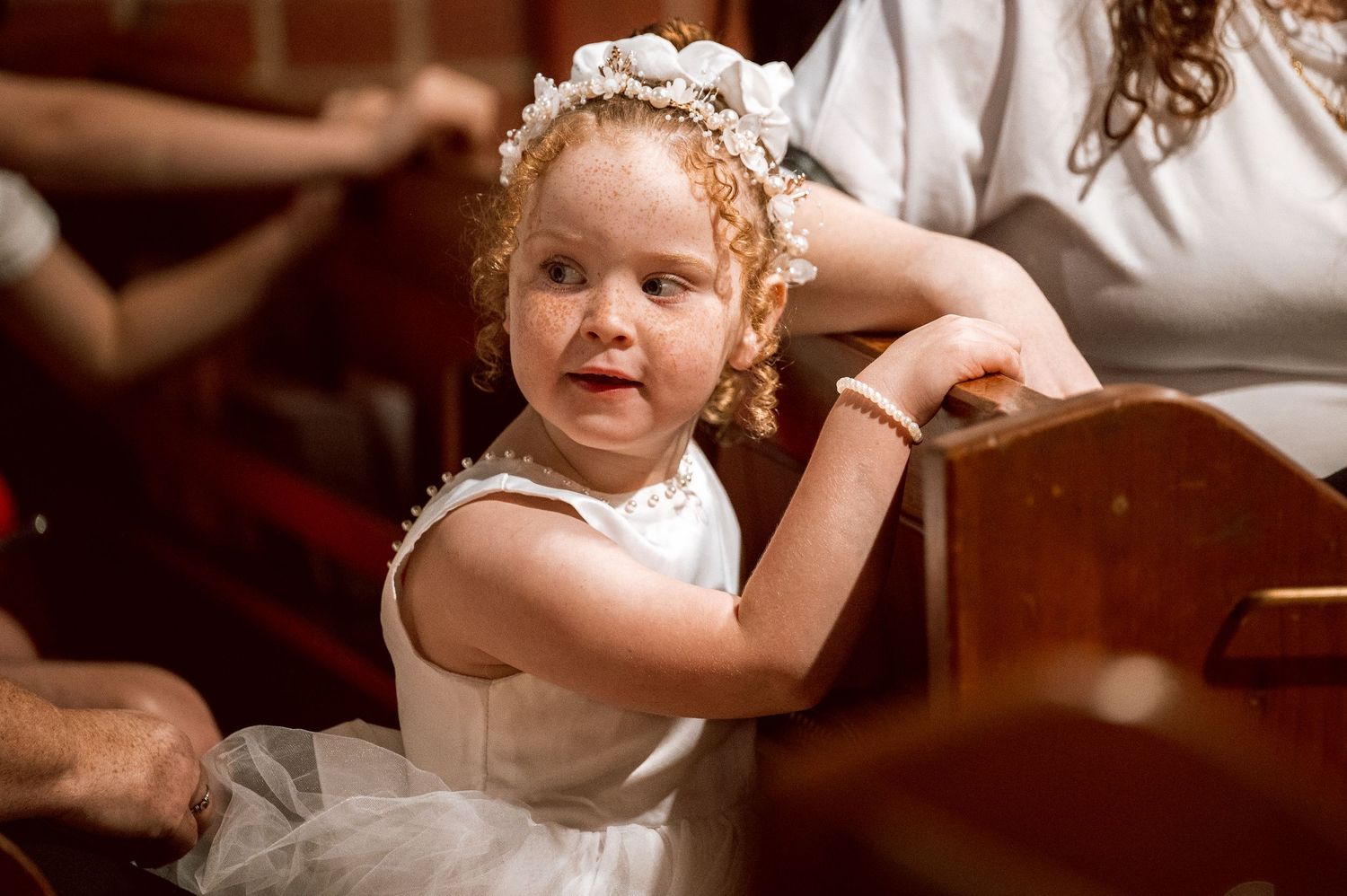 A young child in a white dress and floral headpiece makes an excited expression.