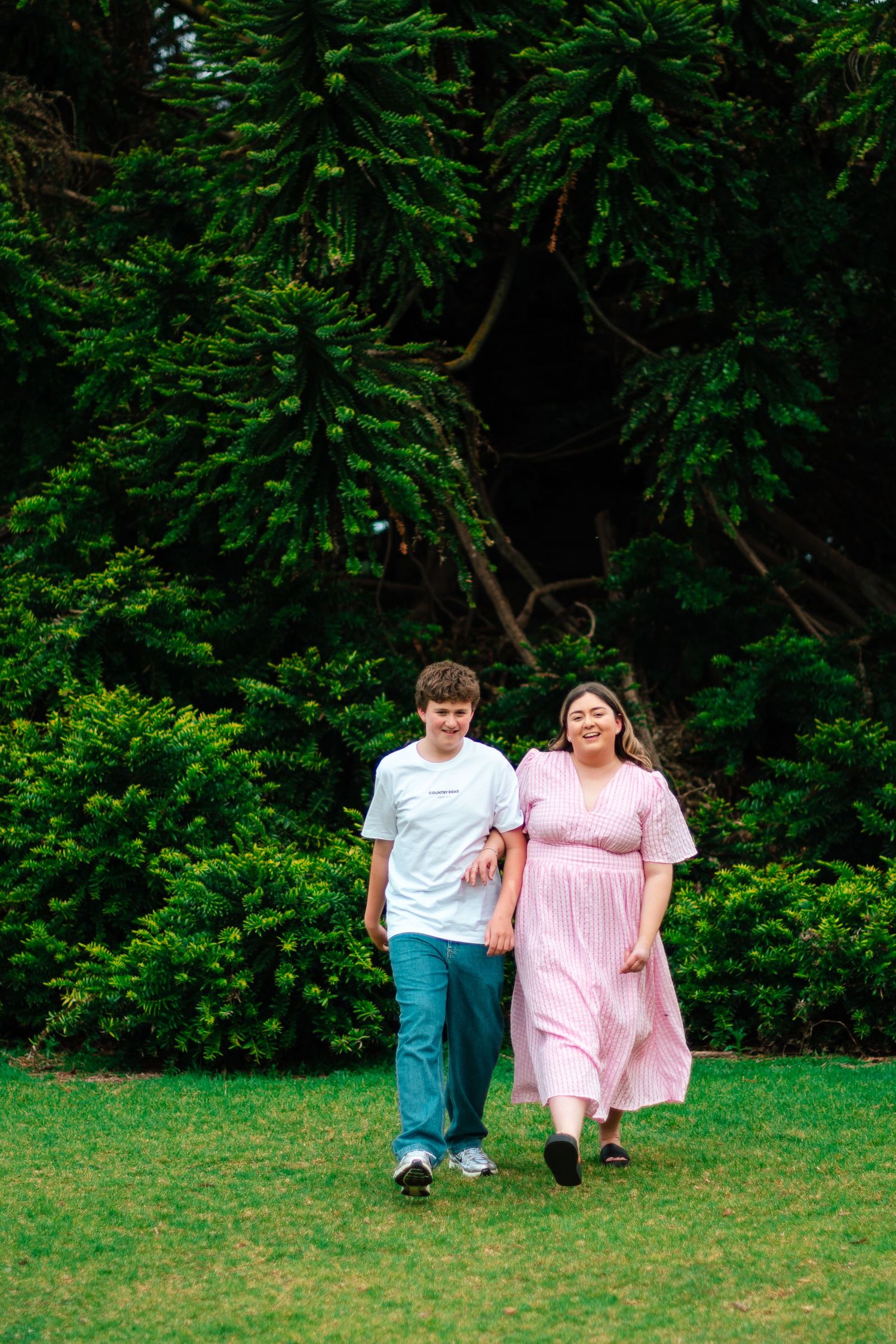 Two people walking together on a grassy lawn surrounded by green foliage.