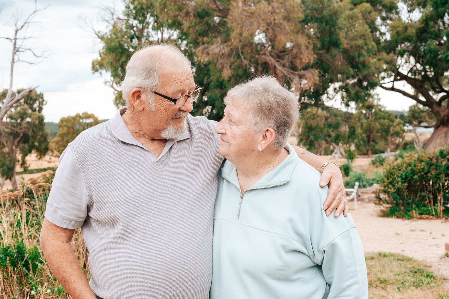 An elderly couple embraces while sharing a tender moment together in a garden setting on a sunny day.