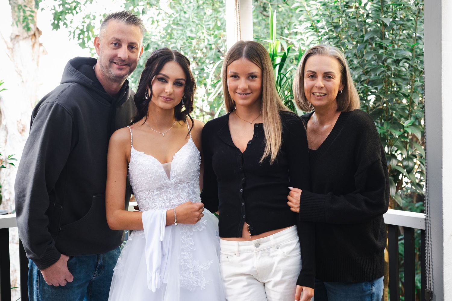 Four people posing together indoors, including a young woman in a white bridal gown, smiling for a photo.