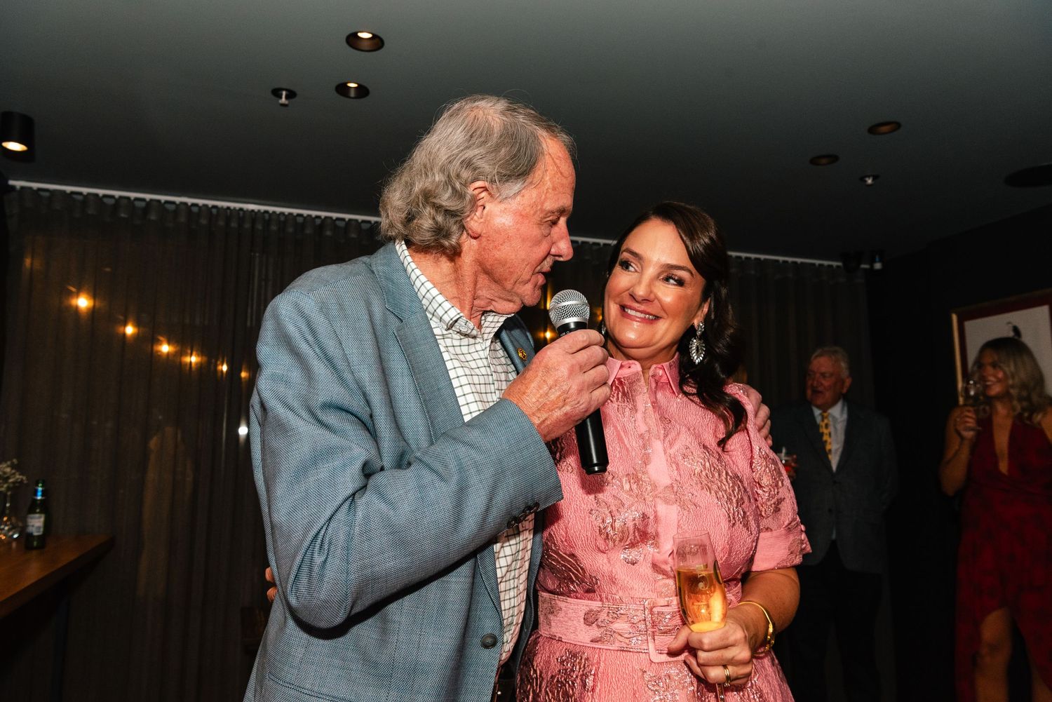 Elderly man speaking into microphone beside smiling woman in pink dress at elegant indoor social event.