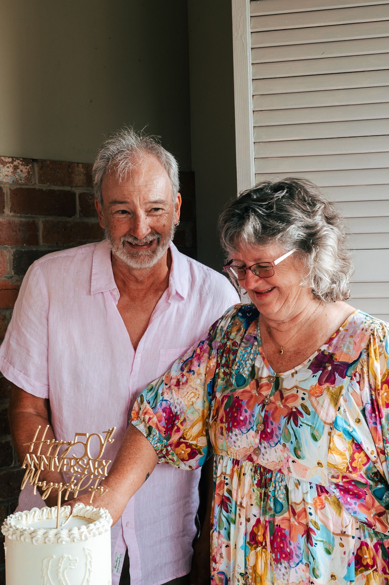 An older couple stands together smiling by a white cake with an anniversary topper at a celebration.