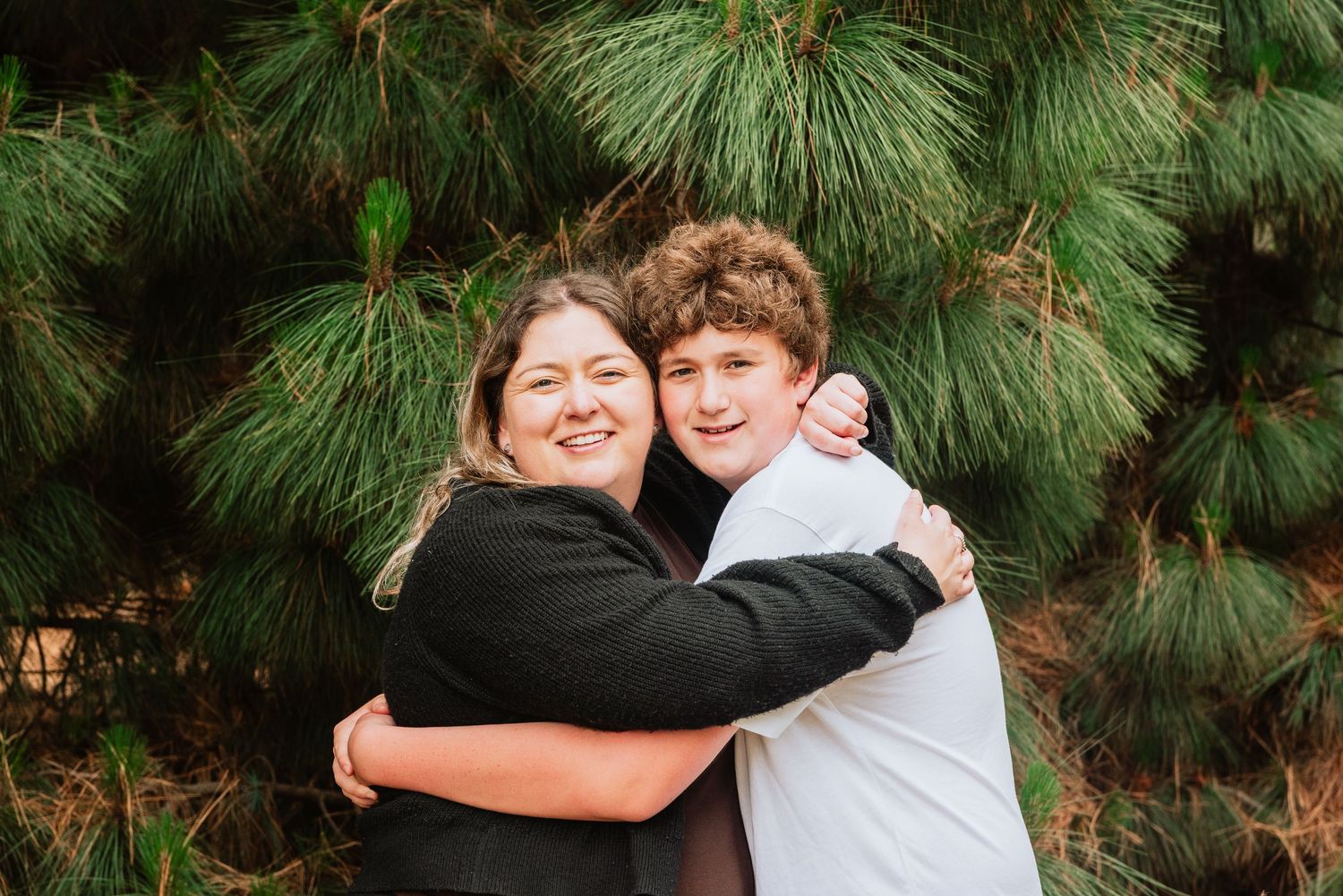 People embracing and smiling outdoors with pine trees in the background.