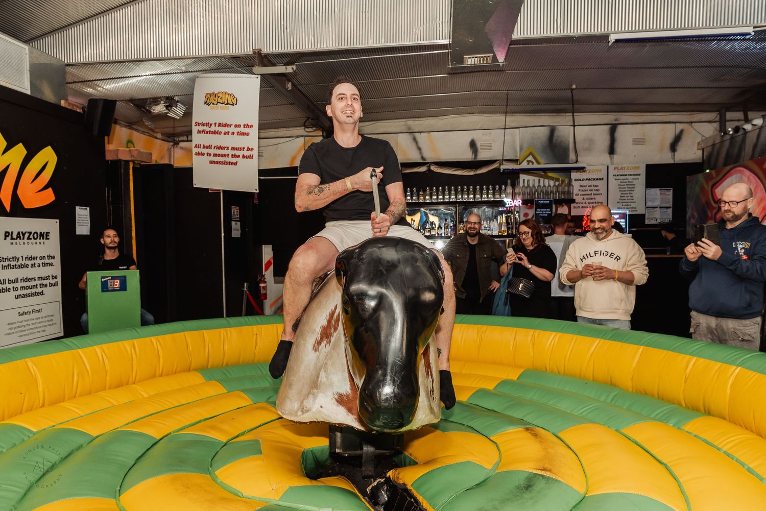 Person riding a mechanical bull at an indoor party venue with spectators watching and cheering in the background.