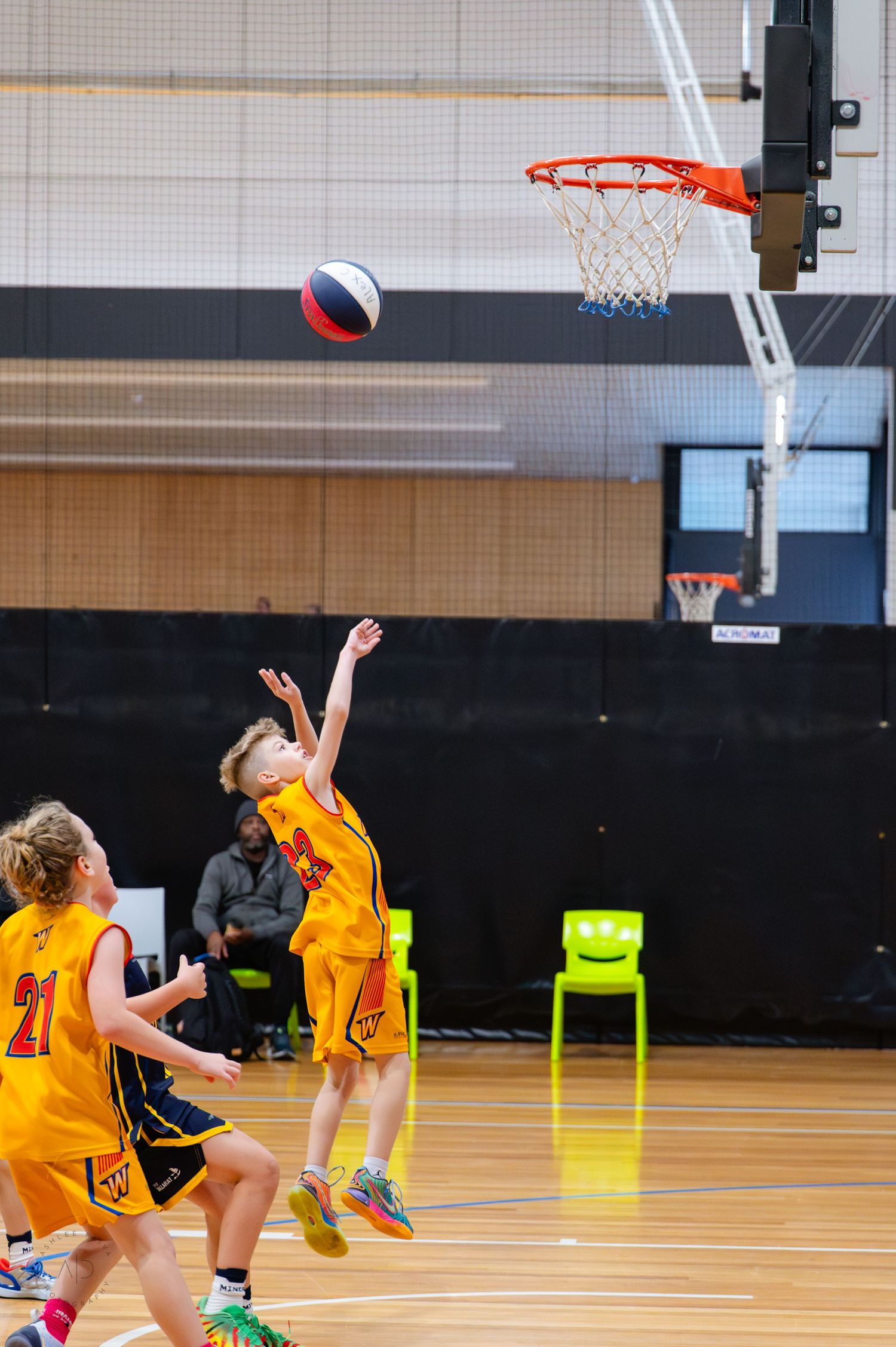 Young basketball players in yellow uniforms compete during an indoor youth basketball game, with one player shooting toward the hoop.