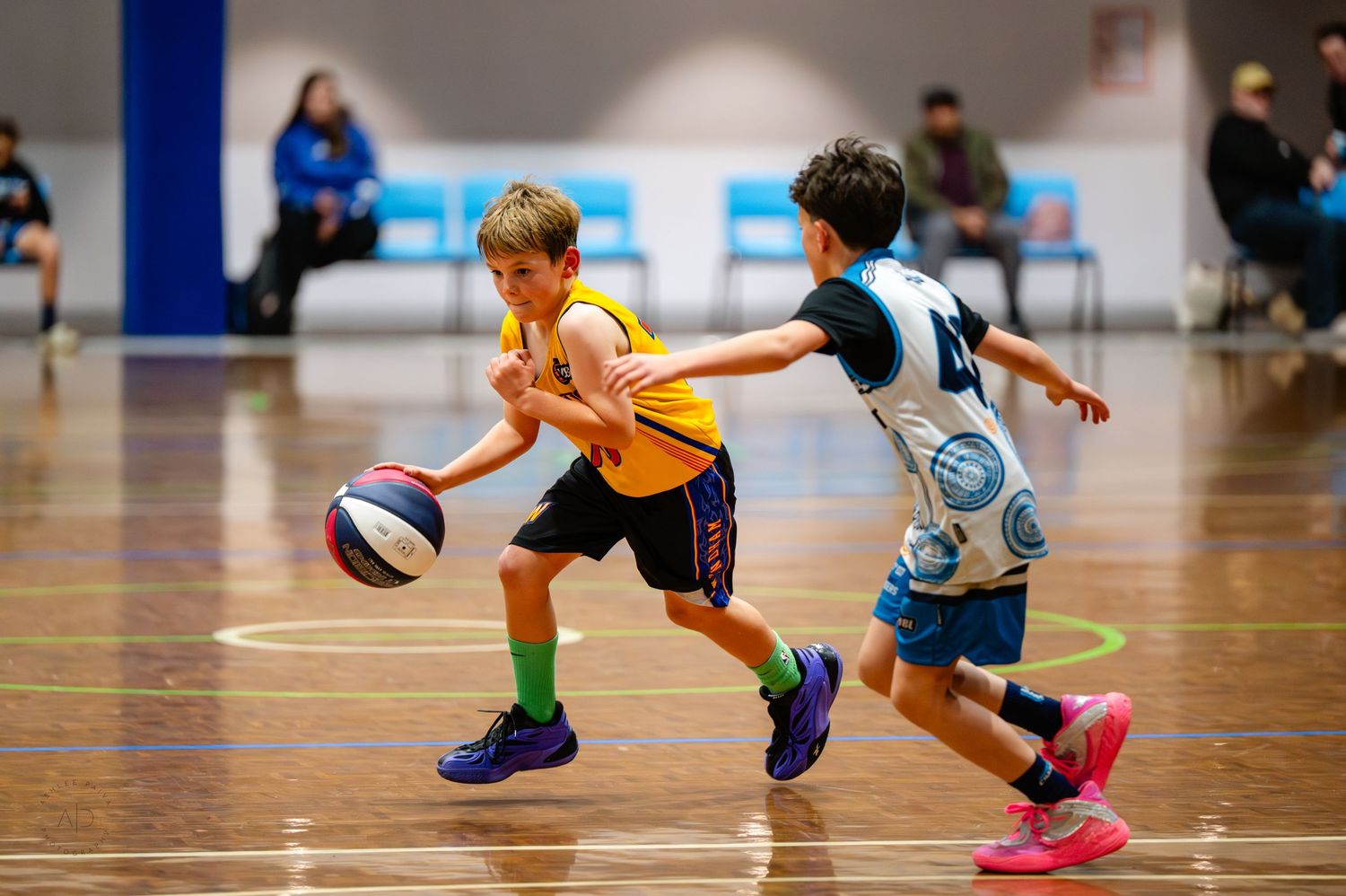 Two young boys playing basketball indoors, one dribbling in an orange jersey while the other defends in a blue uniform.