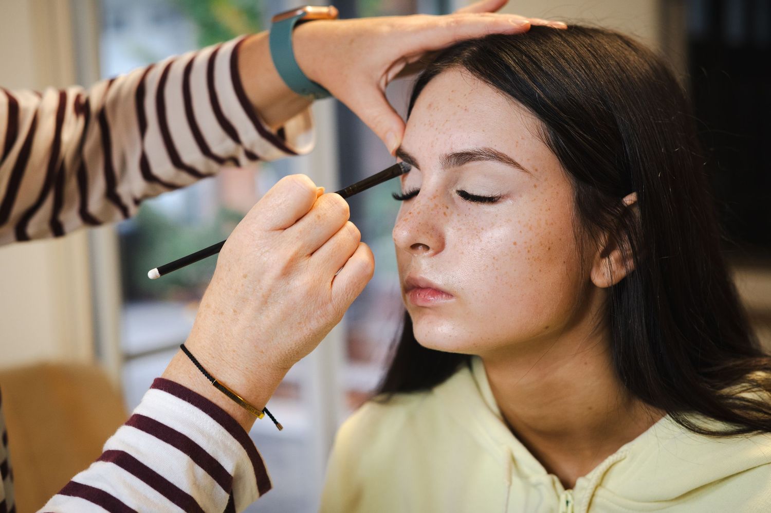 A makeup artist applying eyeshadow to a young woman with freckles wearing a yellow hoodie.