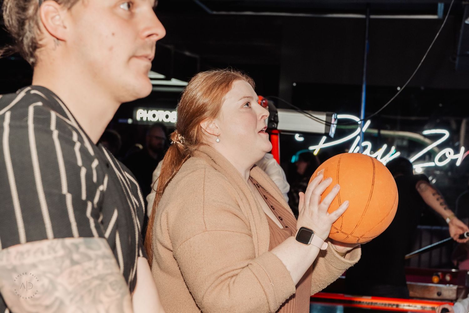 Woman in beige blazer holds orange basketball at indoor arcade venue with neon signs in background.