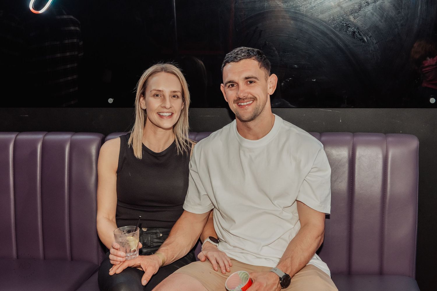 A smiling couple sitting together on a purple booth seat in a nightclub, woman holding a drink.