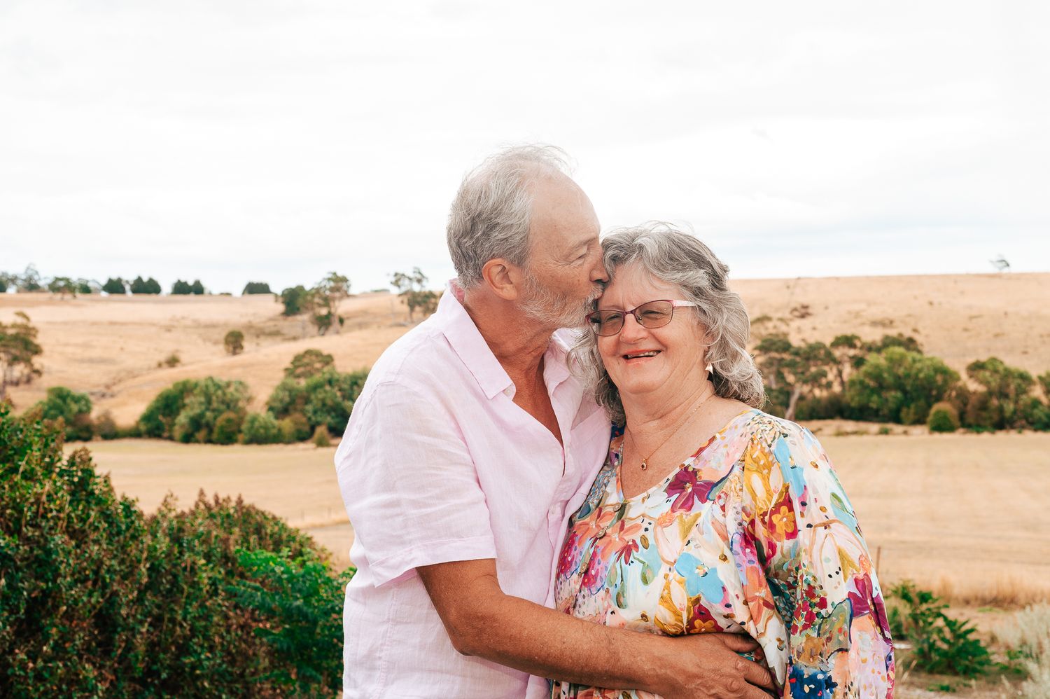 Elderly couple embraces while standing in front of a rural landscape with rolling hills and scattered trees.
