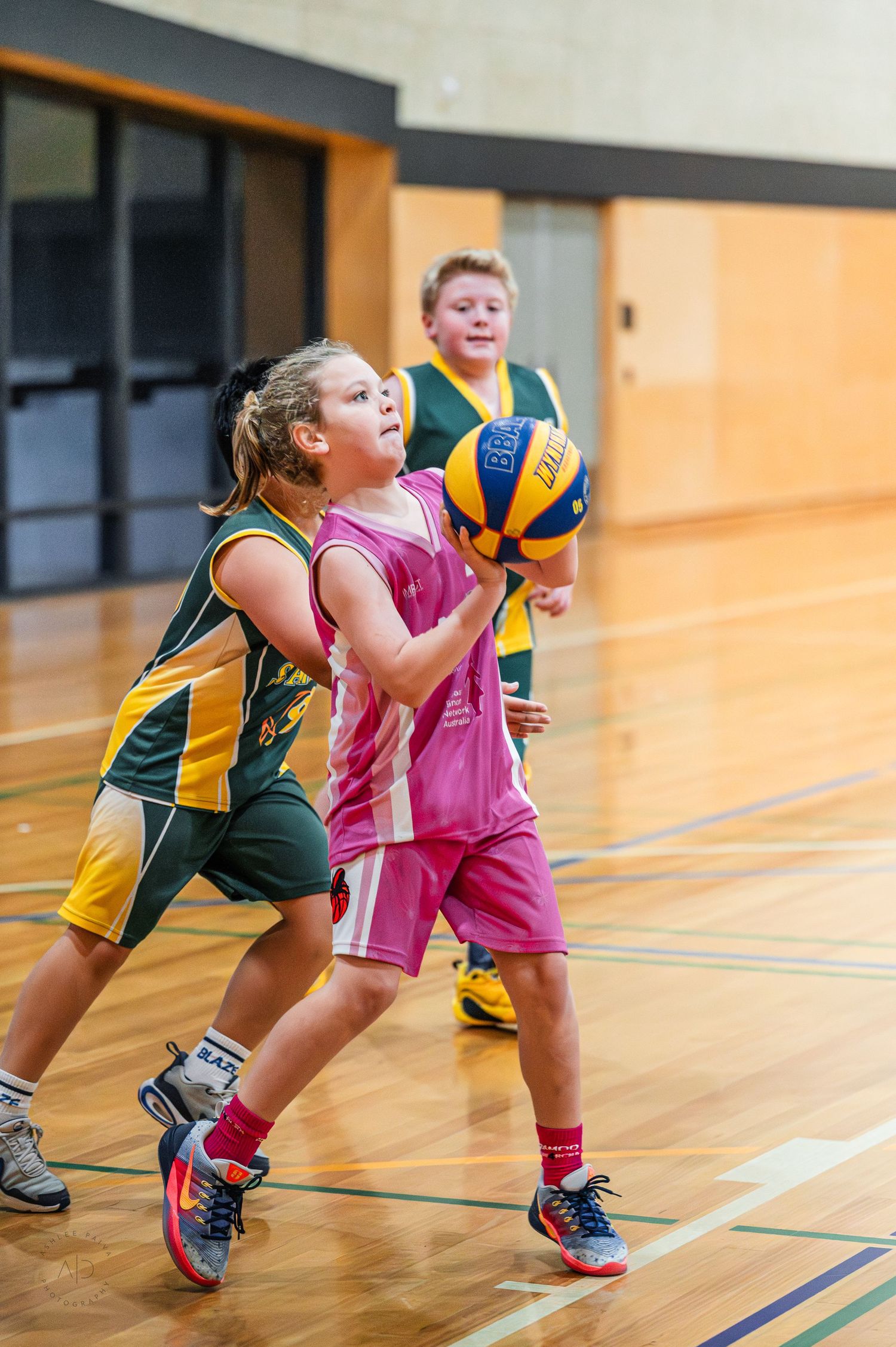 Young girls playing basketball in indoor court, one in pink uniform shooting while defender in green watches.