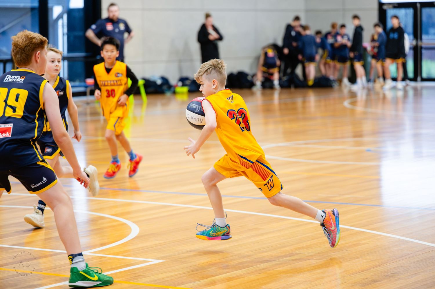 Young boys playing competitive indoor basketball game, player in yellow jersey dribbling ball on hardwood court.