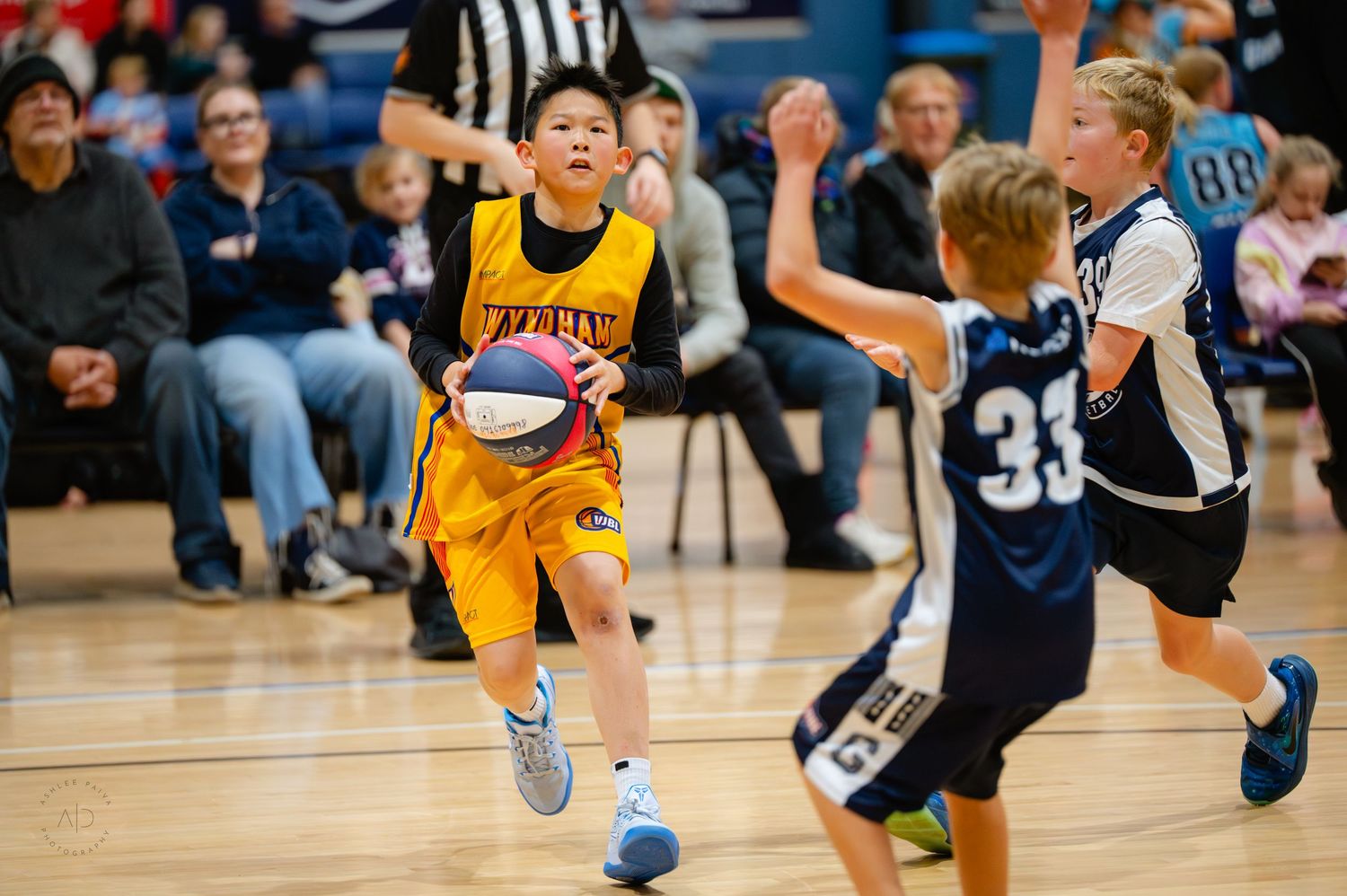 Young basketball players compete in a youth league game, with a player in yellow dribbling past defenders in blue uniforms.