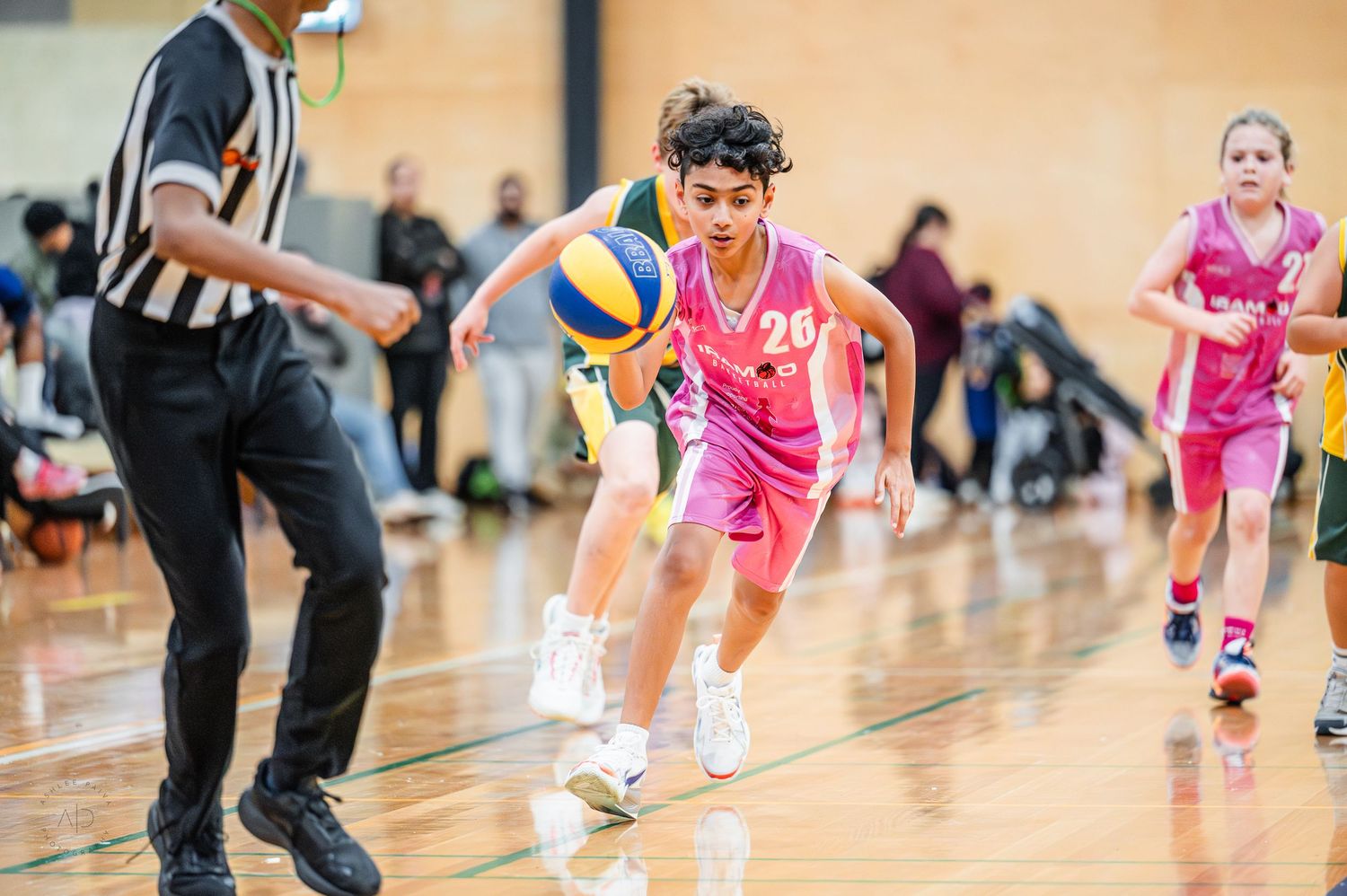 Young basketball player in pink jersey dribbling during a youth indoor game with referee nearby.