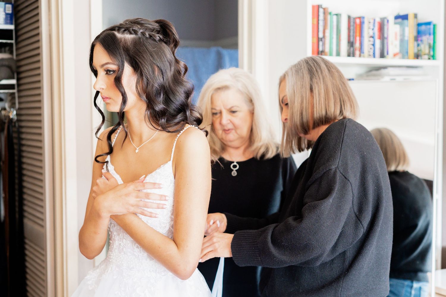 Bride in white lace gown being helped by two women while getting ready for her wedding day.