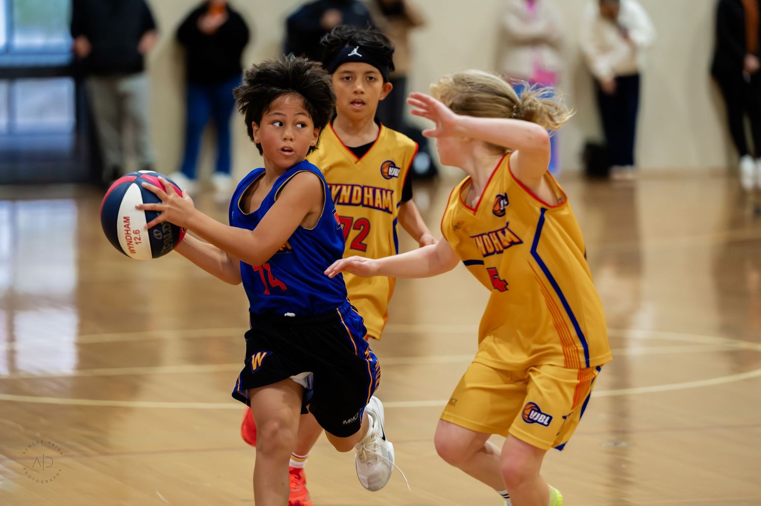 Young basketball players compete in an indoor game, with a player in blue driving past defenders in yellow Wyndham uniforms.