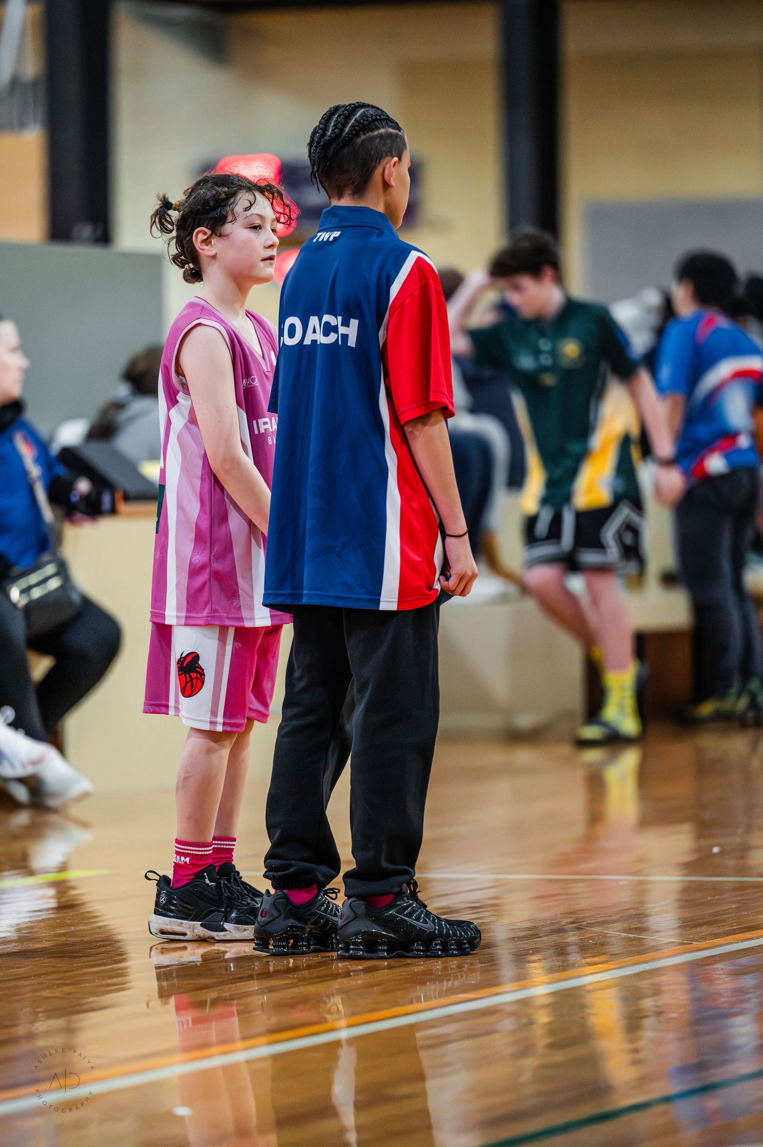 Two young basketball players stand on an indoor court during a youth basketball game in a gymnasium.