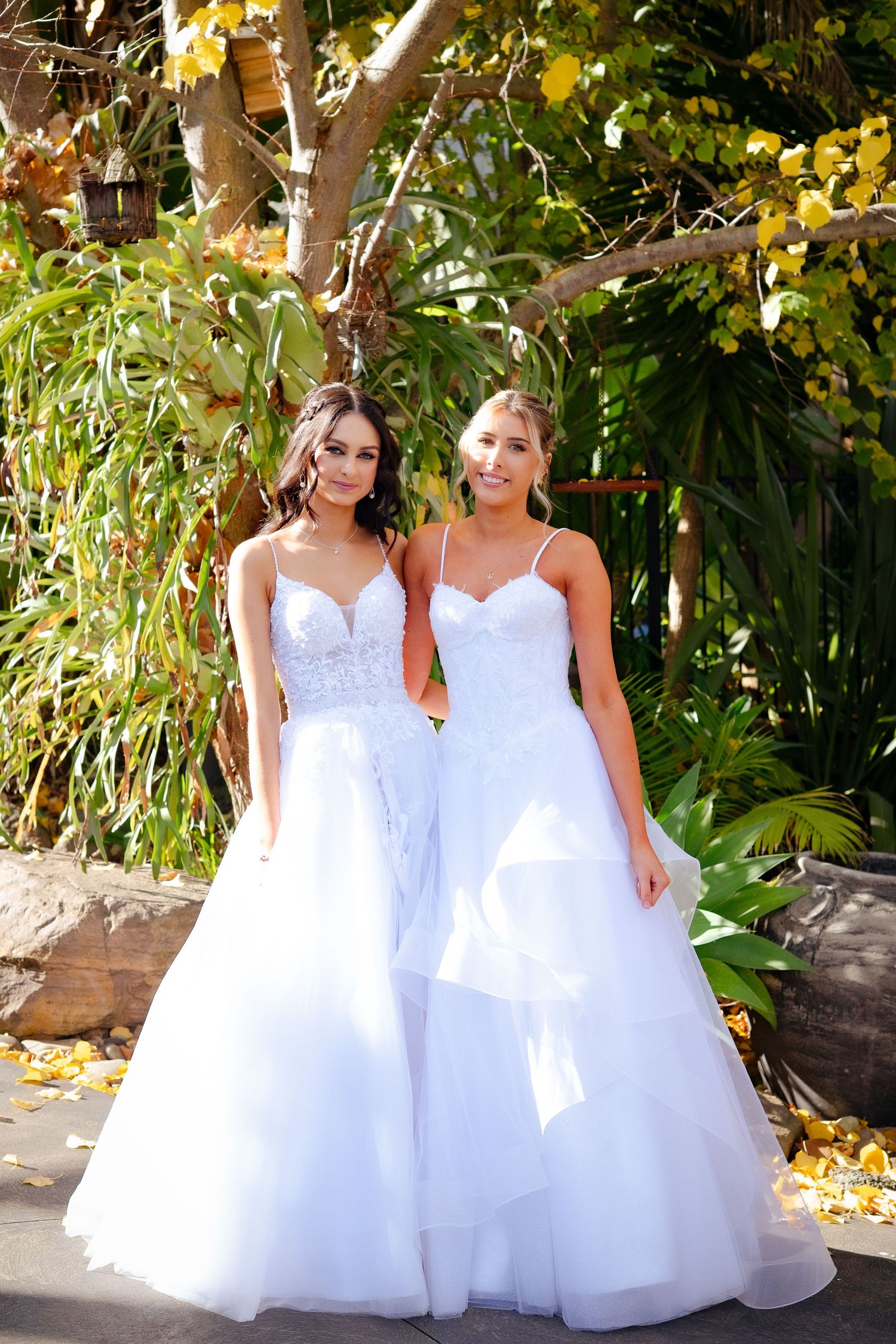 Two brides in white ball gowns with lace bodices pose outdoors among lush green trees and yellow flowers.