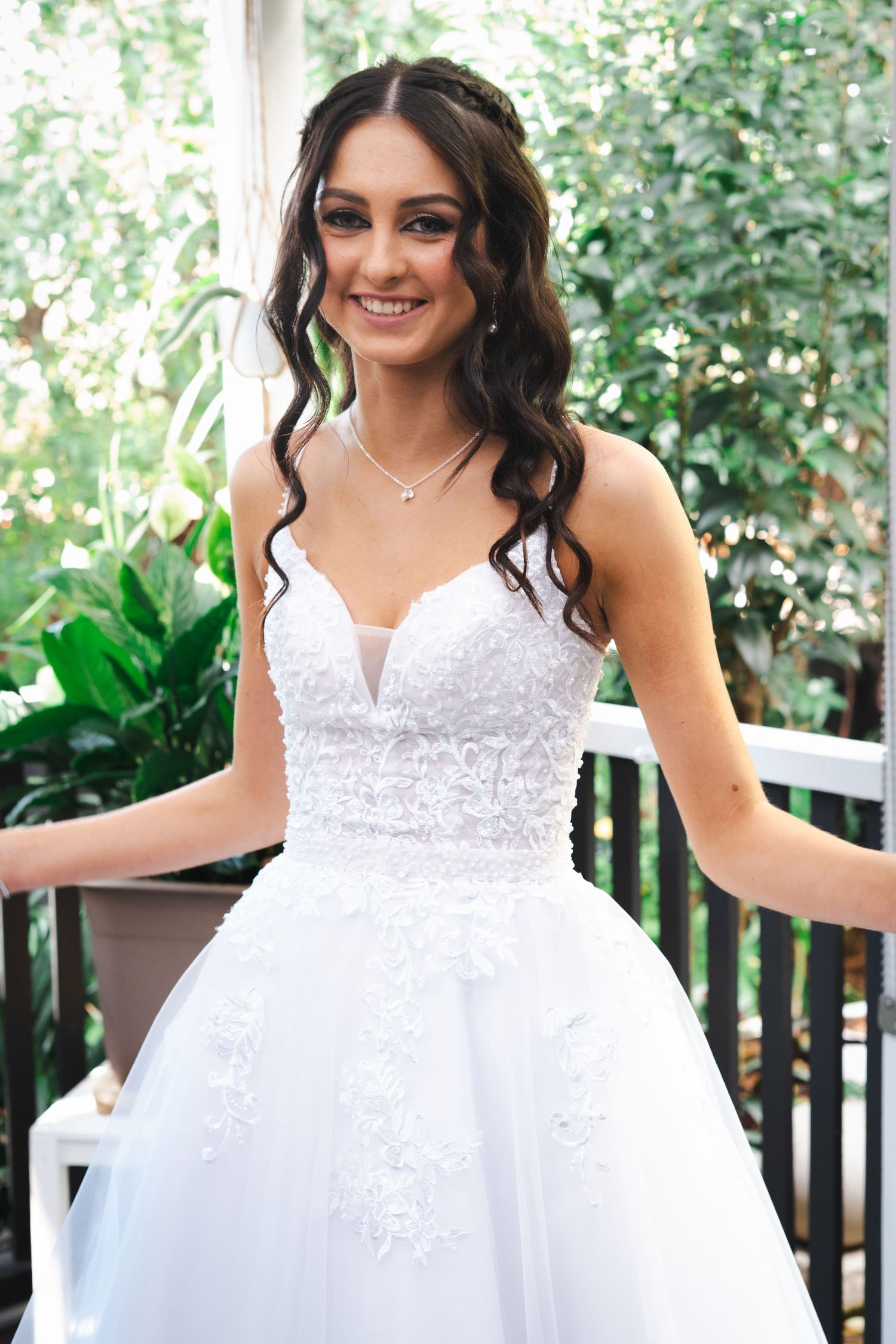 Smiling bride in white lace ball gown wedding dress with spaghetti straps, floral appliques, and delicate necklace outdoors.