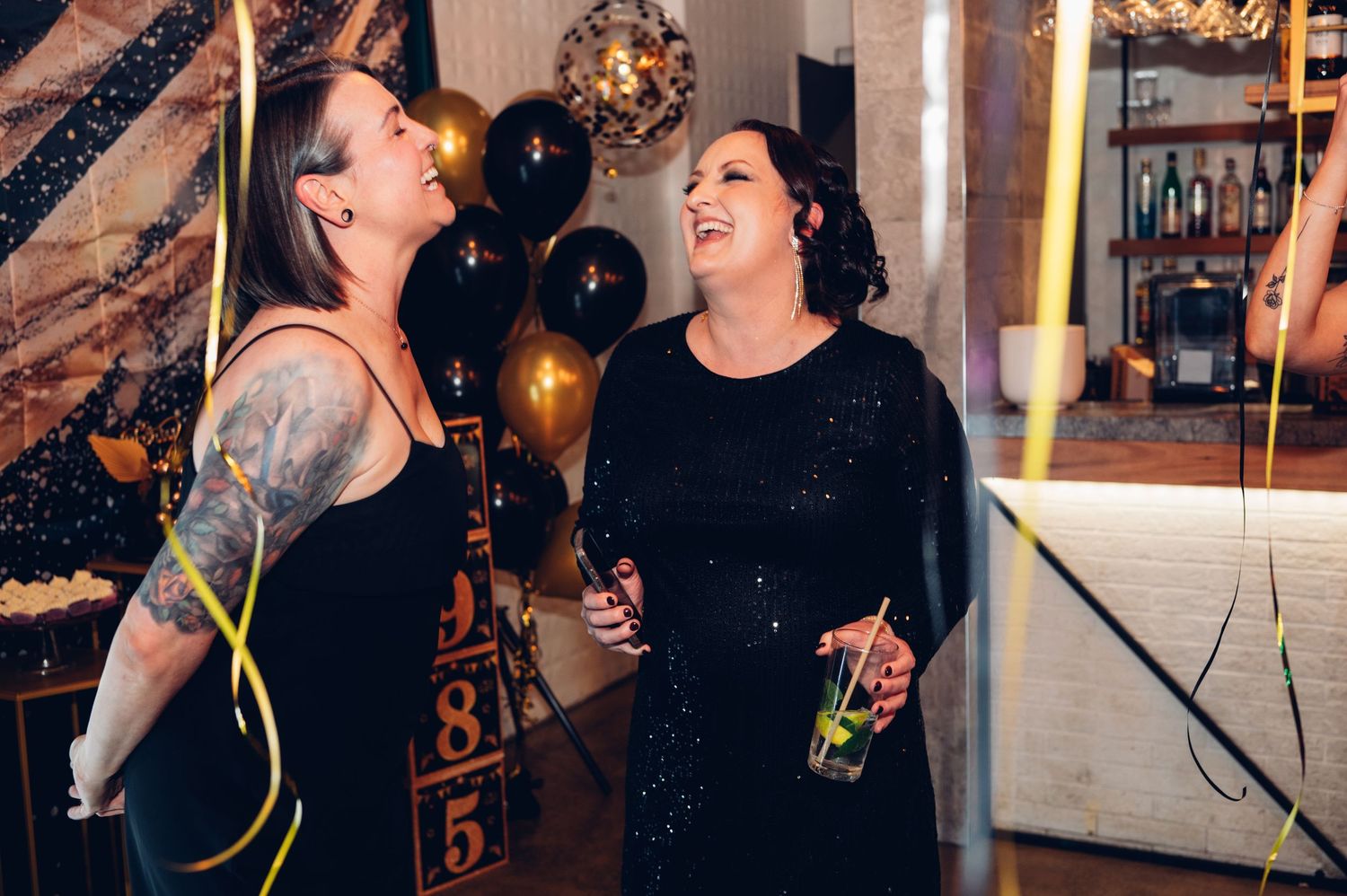 Two people sharing a laugh at a party decorated with black balloons and festive lighting.