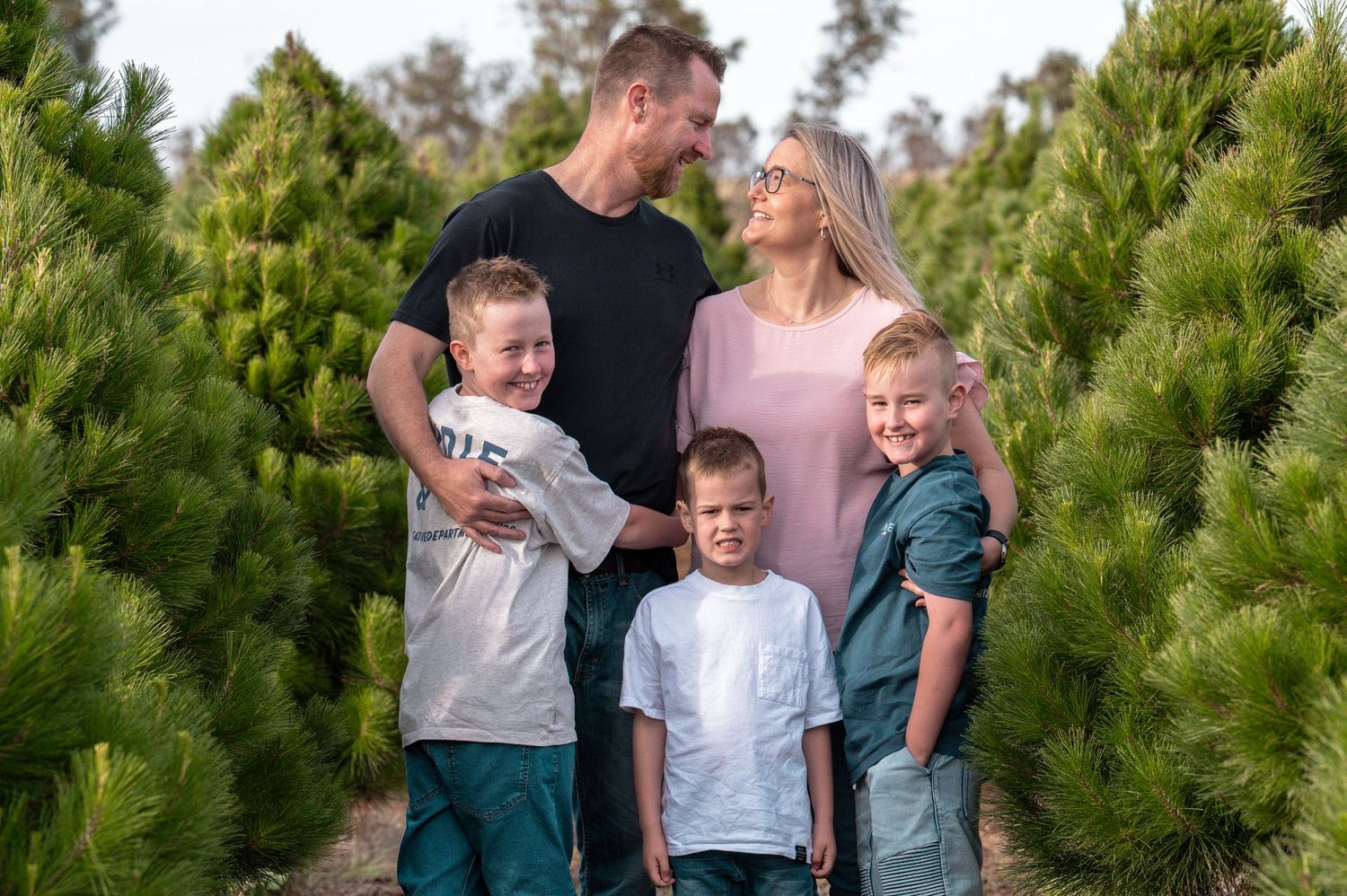 Family posing together outdoors among evergreen trees at a Christmas tree farm.