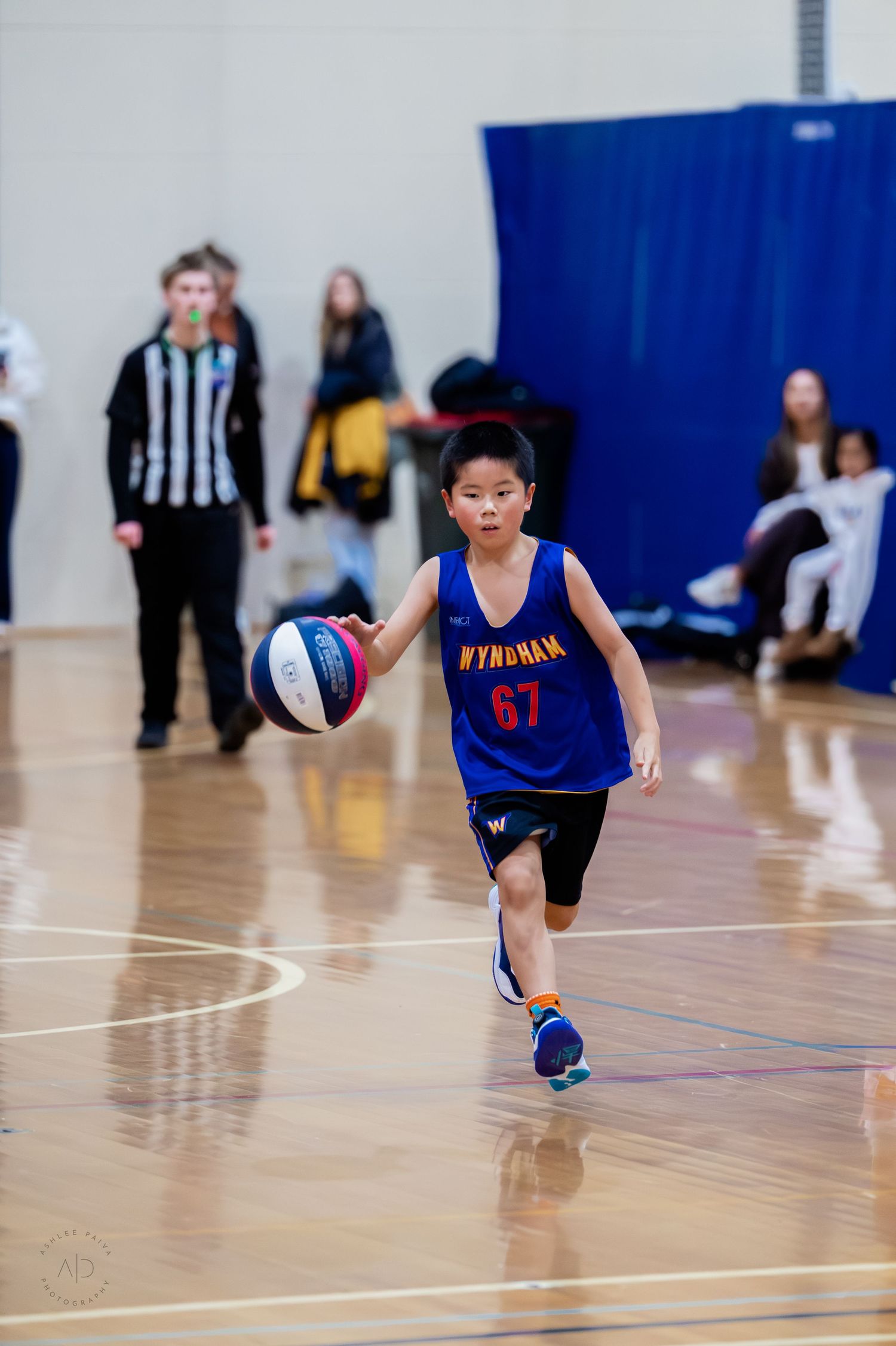 Young boy in Wyndham #67 blue jersey dribbling basketball on indoor court during youth game.