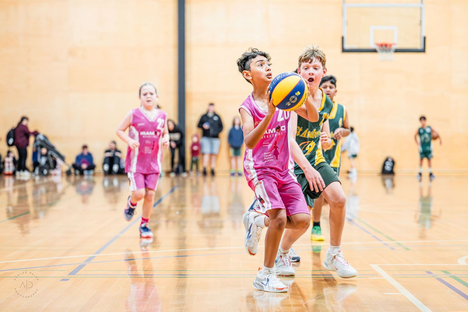 Young basketball players compete in an indoor court game, with a player in pink uniform reaching for the ball.