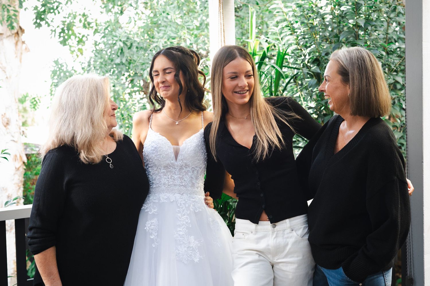 Bride in white lace gown smiling with three women in black tops during wedding preparation indoors.