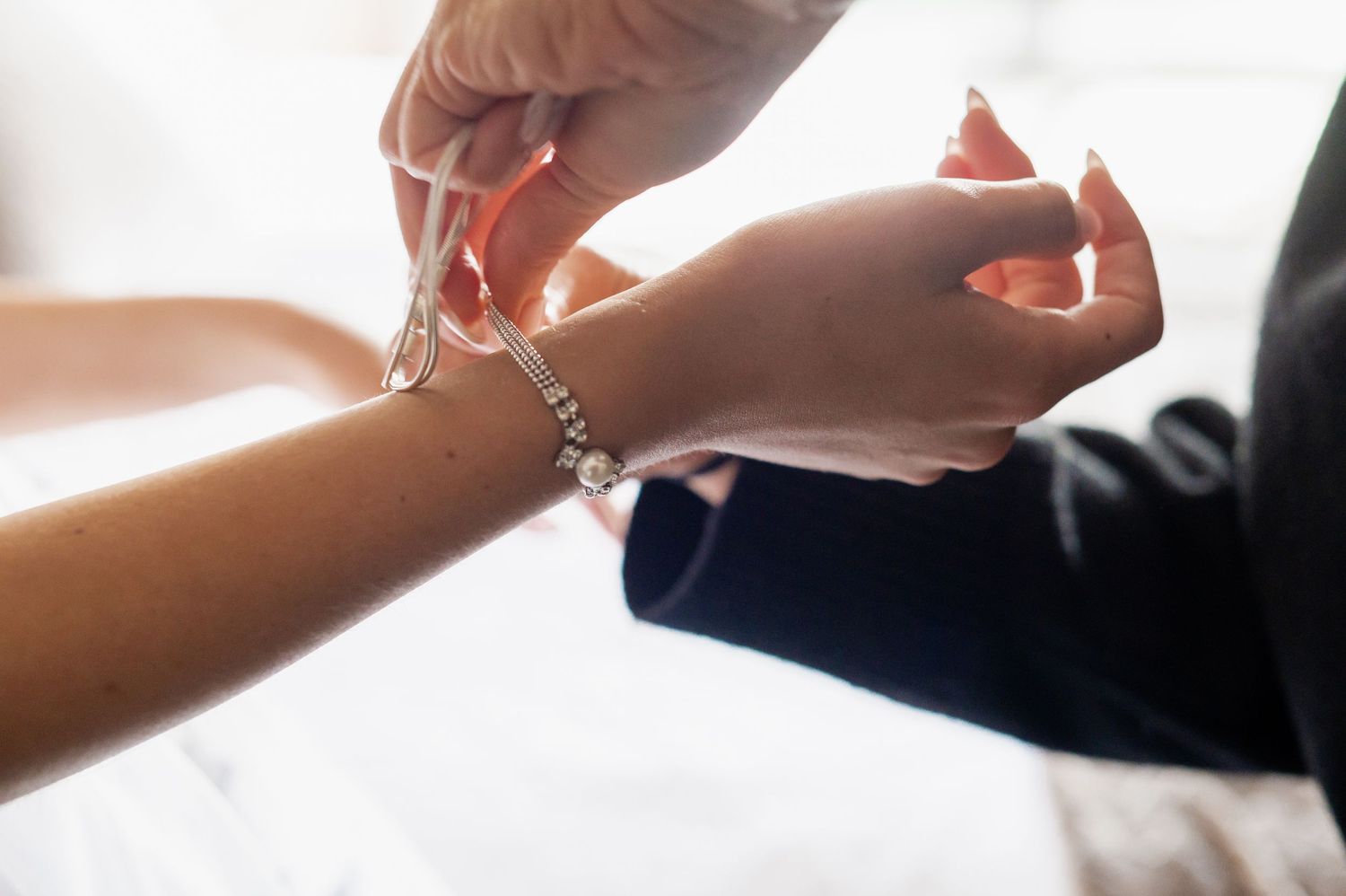 Close-up of hands fastening a delicate pearl and crystal bracelet on a bride's wrist at a wedding.