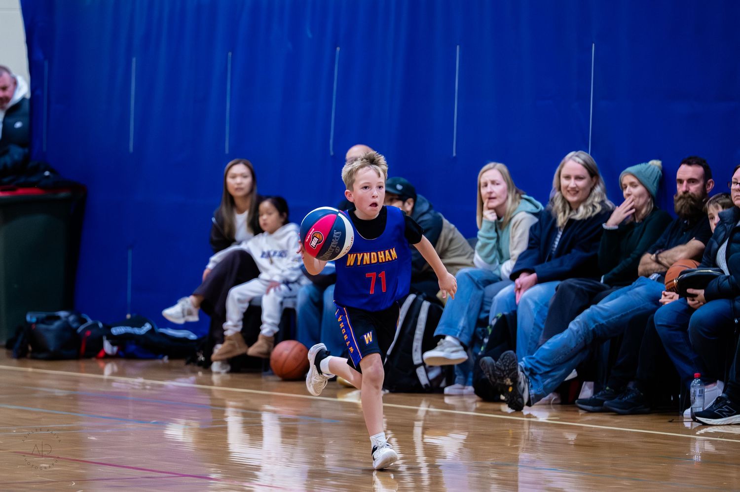 Young basketball player in blue Strahan jersey dribbling down court during indoor youth game with spectators watching.