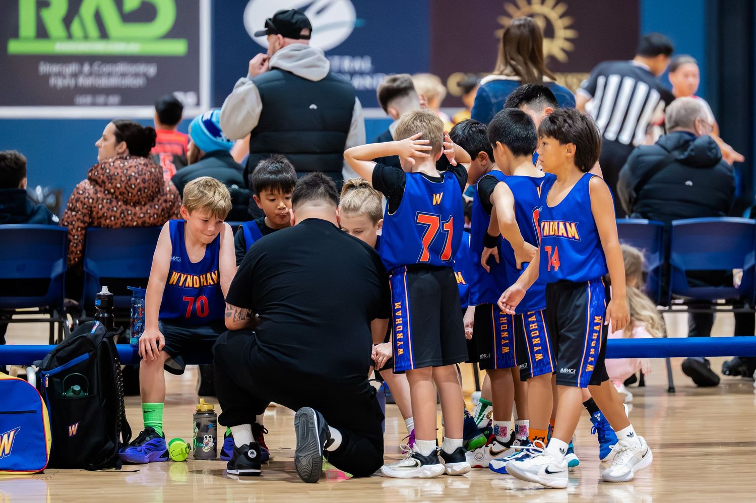 Youth basketball team huddles with coach during game, players wearing blue uniforms numbered 70 and 71 on indoor court.