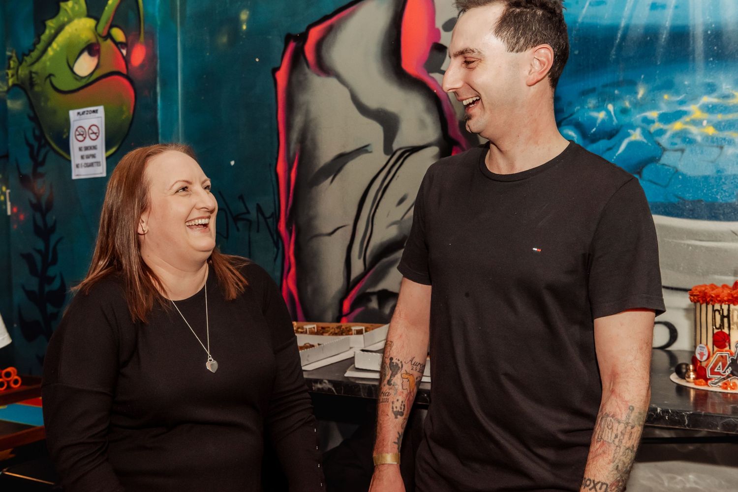 A smiling man and woman in black shirts laugh together near colorful graffiti wall art at a casual event.
